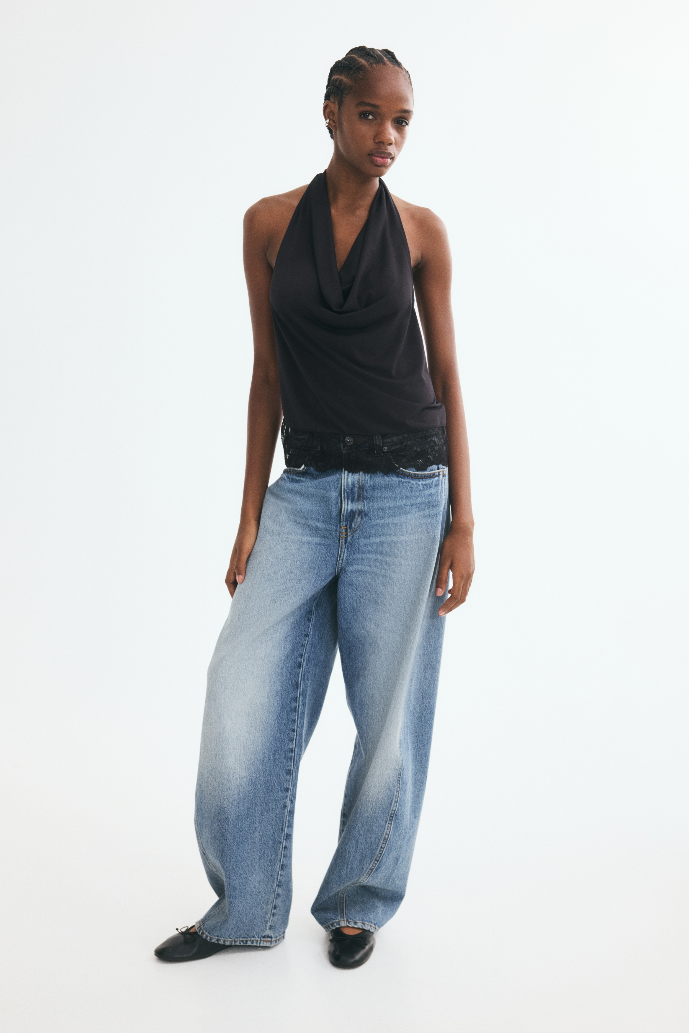 Facing the front, a black halter top features a draped cowl neckline and a relaxed silhouette. The sleeveless top features a wide, intricate lace trim along the straight hemline.