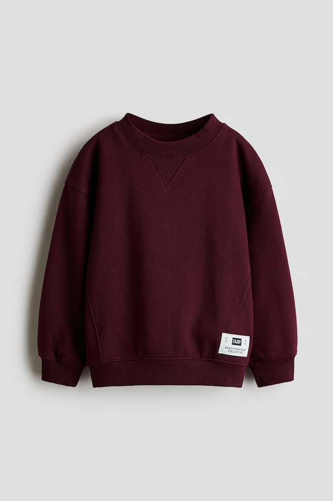A burgundy cotton sweatshirt is facing front, showcasing a ribbed crew neck with a V-insert detail and dropped shoulders. It is displaying ribbed cuffs, a ribbed hem, and a small white rectangular label near the bottom right hem. - 1