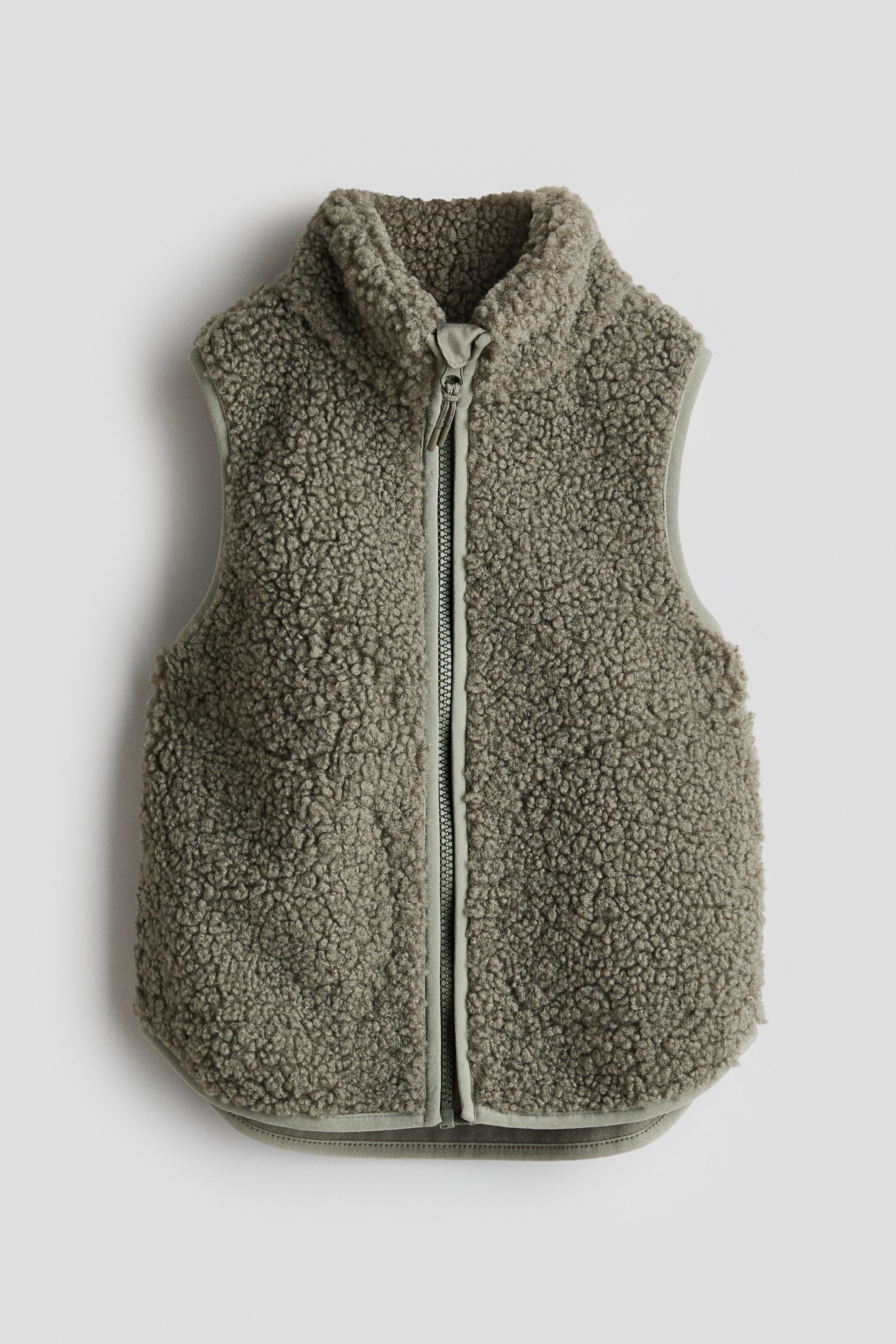 View larger image: A muted olive green teddy fleece vest is showcasing its front, featuring a full-length zip with a fabric pull and a standing collar. The vest is completed with smooth binding along the armholes and a rounded bottom hem.