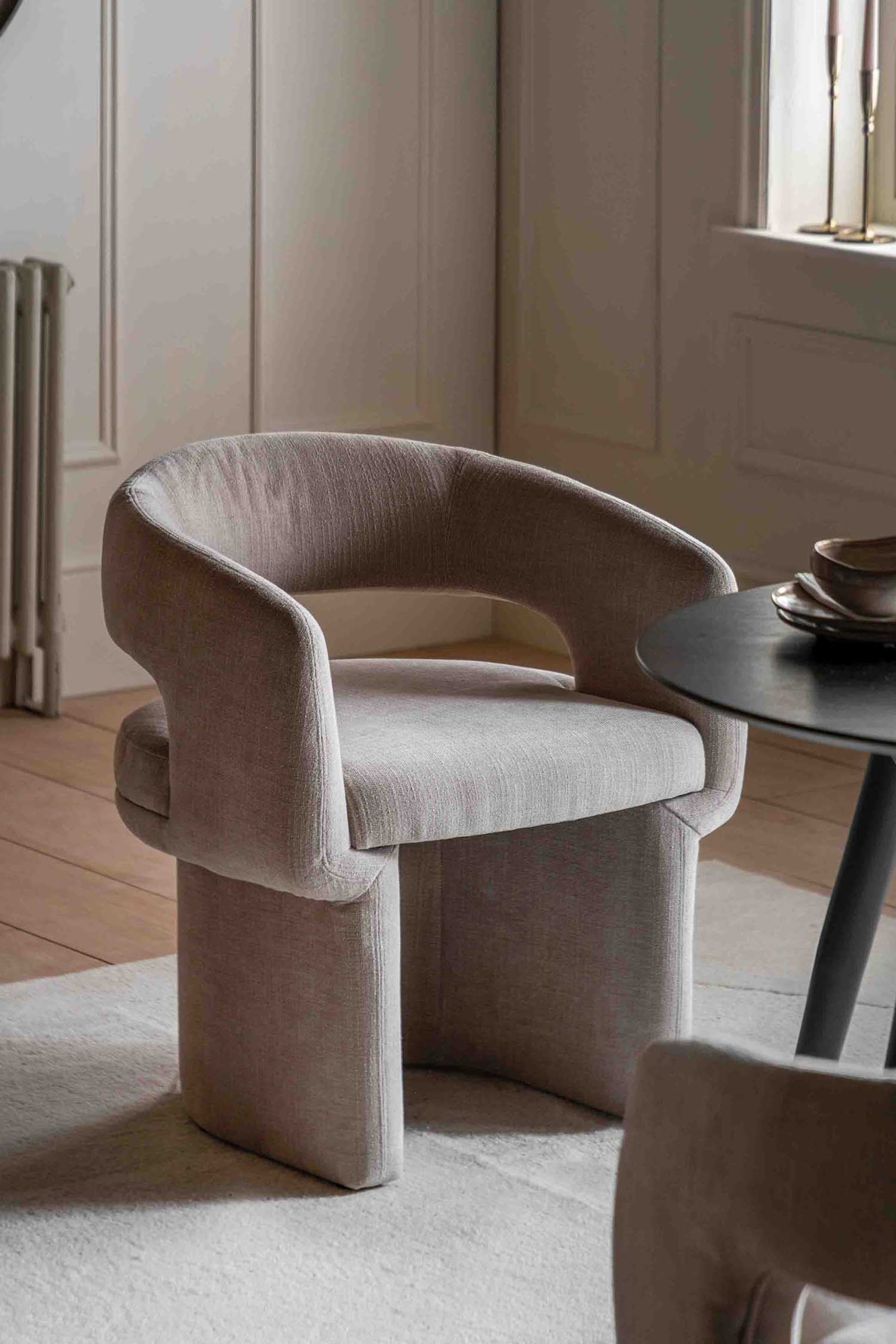 View larger image: A light beige upholstered chair is showcasing its front-right side, featuring a curved backrest and integrated armrests with side cut-outs. The chair is designed with a solid, upholstered base formed by two wide, curved supports.