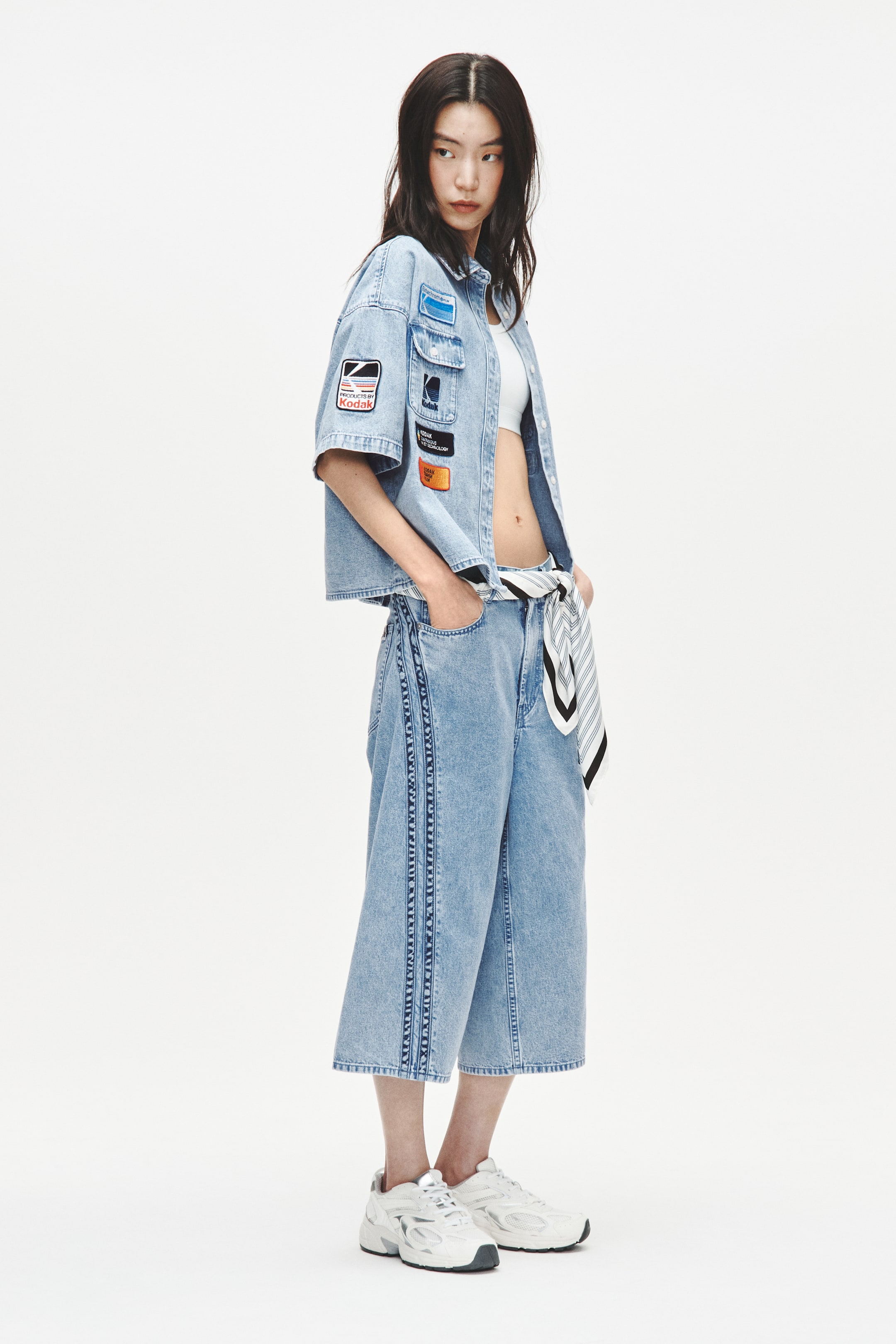 Xem ảnh lớn hơn: Một bộ trang phục denim màu sáng đang được giới thiệu, bao gồm áo sơ mi ngắn tay với nhiều miếng vá ở phía trước và tay áo, không cài cúc bên ngoài áo crop top màu trắng. Bộ trang phục này bao gồm quần ống rộng lửng với các sọc dọc denim tối màu, khăn quàng cổ sọc đen và trắng buộc ở eo, và giày thể thao chunky màu trắng và xám.