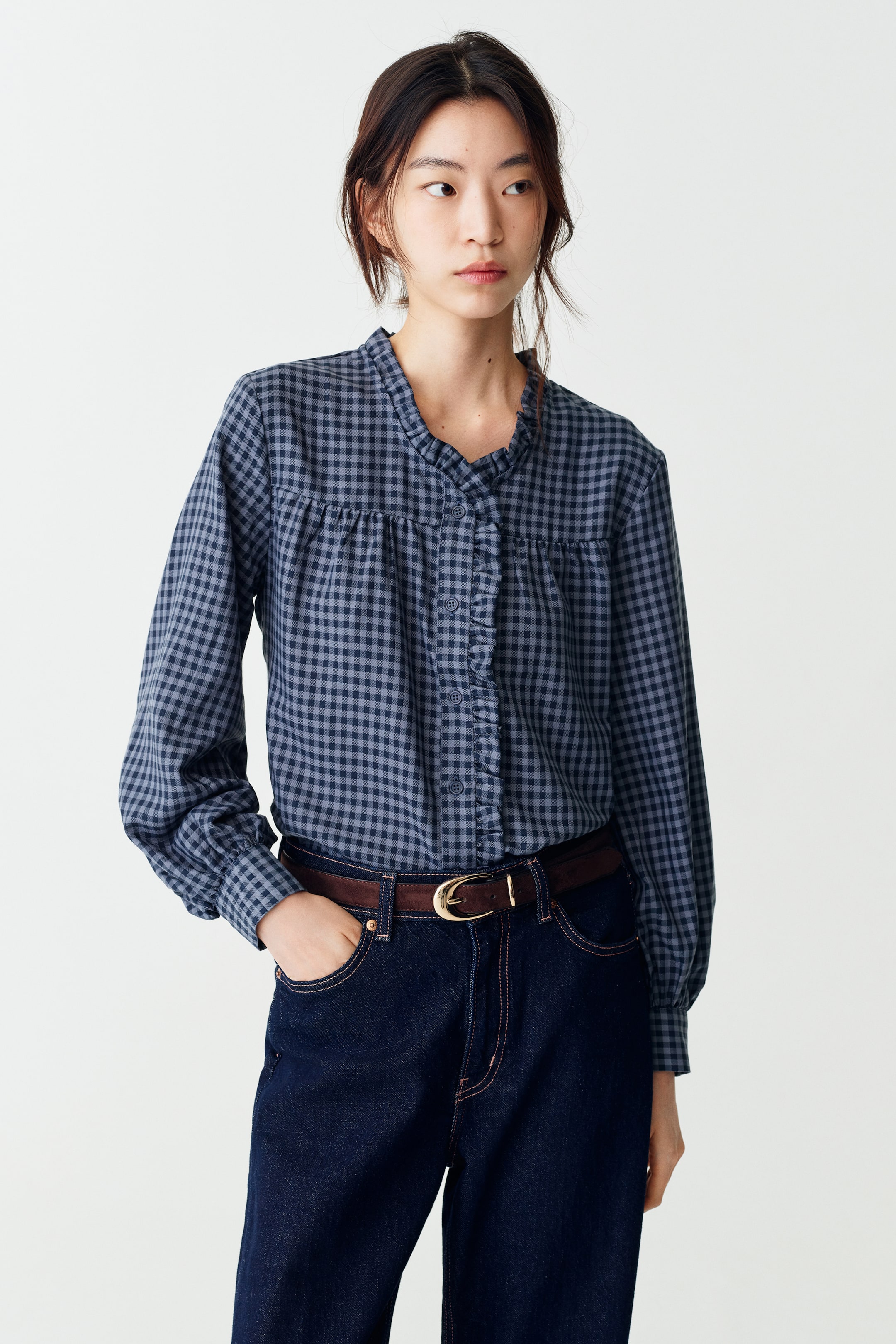 Xem ảnh lớn hơn: Người mẫu đang mặc một chiếc áo blouse vải twill họa tiết gingham, nổi bật với cổ áo nhỏ có bèo nhún, đường viền bèo nhún dọc phần thân trước cài nút, cầu ngực nhún, và tay dài với cổ tay nhún. Áo được sơ vin vào quần jeans denim tối màu, phối cùng thắt lưng da nâu.