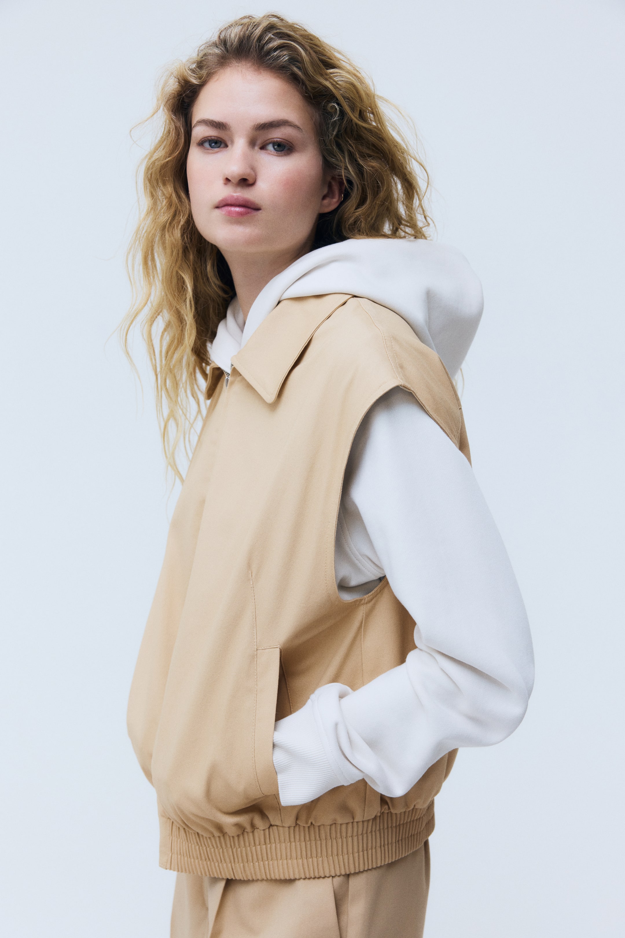 View larger image: A tan sleeveless jacket is shown from a slight side-front angle, displaying a pointed collar and a partial front zip. It features side welt pockets and an elasticated ribbed hem, and is layered over a white hooded sweatshirt.