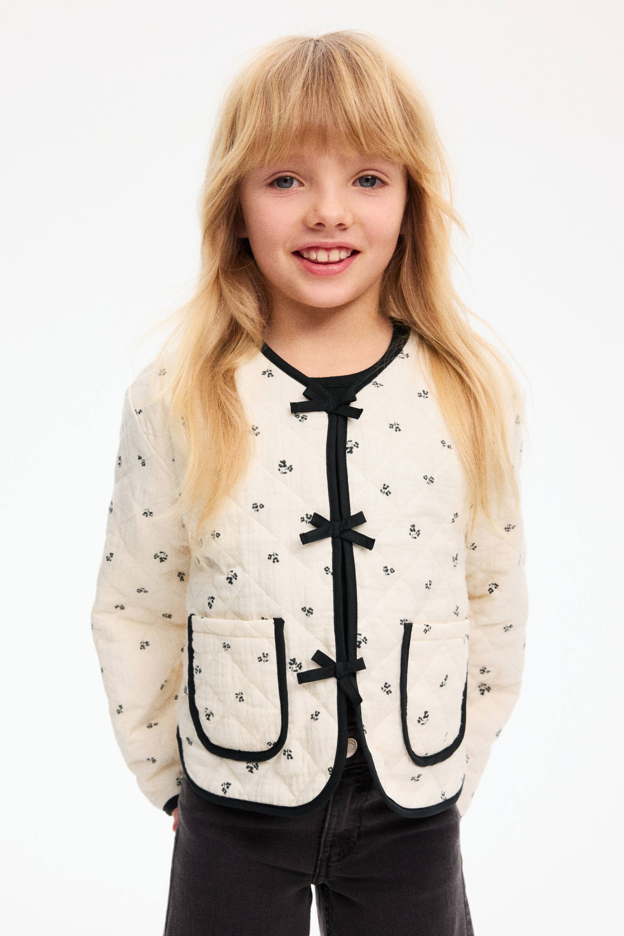 Ampliar la imagen: La chaqueta acolchada color crema está de frente, mostrando un pequeño estampado floral oscuro y un acolchado en rombos. Está detallada con ribetes negros a lo largo del escote redondo, la apertura frontal, el dobladillo y dos bolsillos tipo parche frontales, y se cierra con lazos de cinta negros en el centro delantero.