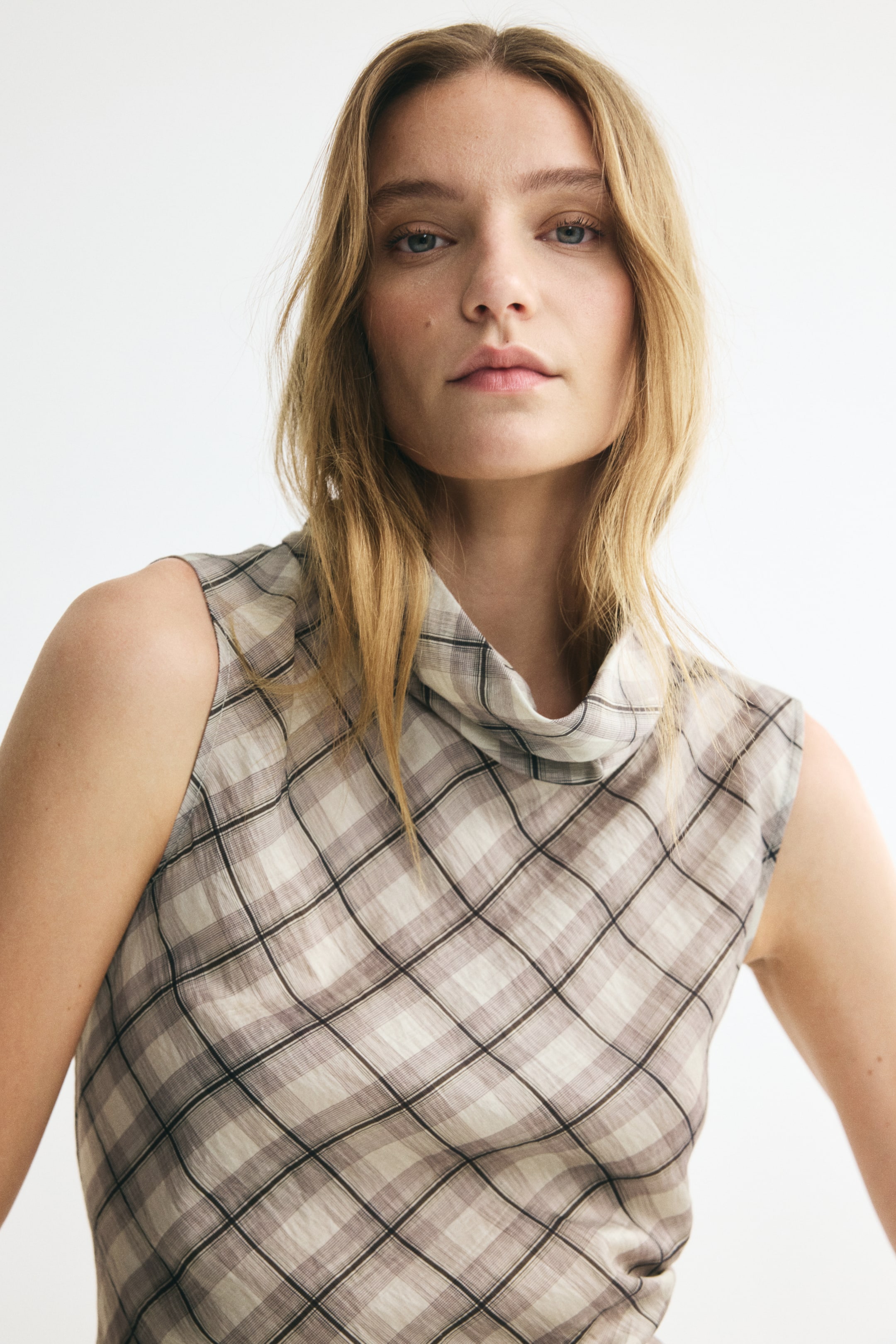 View larger image: A sleeveless top in a neutral-toned plaid pattern is displaying a high, draped turtleneck and a lightweight, textured weave from a front angle. The diagonal check design is showcasing shades of beige, brown, and white.