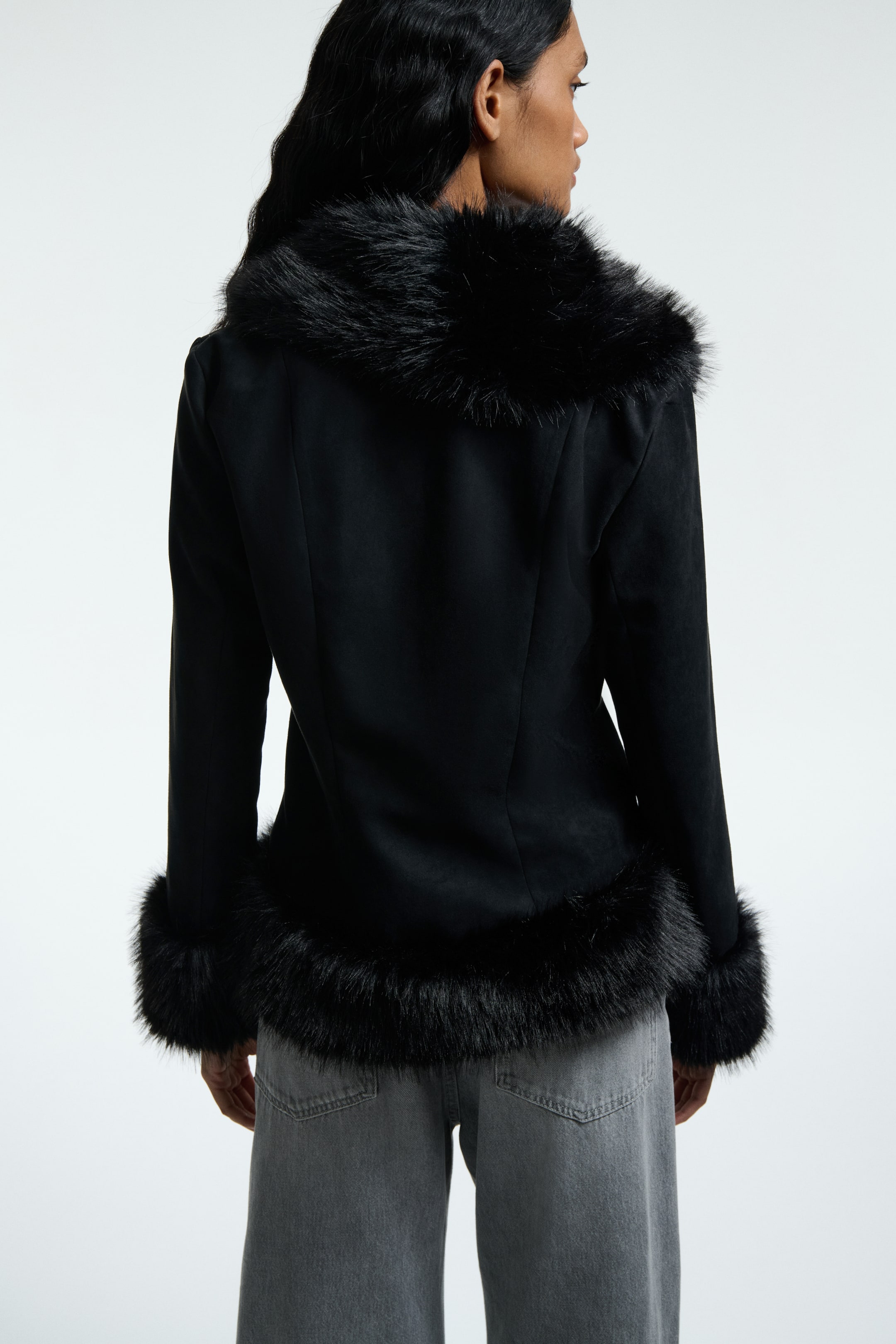 Agrandir l'image: La veste noire est présentée de dos, mettant en valeur un large col en fausse fourrure moelleux. Elle présente également des poignets en fausse fourrure assortis et une garniture en fausse fourrure le long de l'ourlet inférieur, contrastant avec le tissu noir mat de la veste.