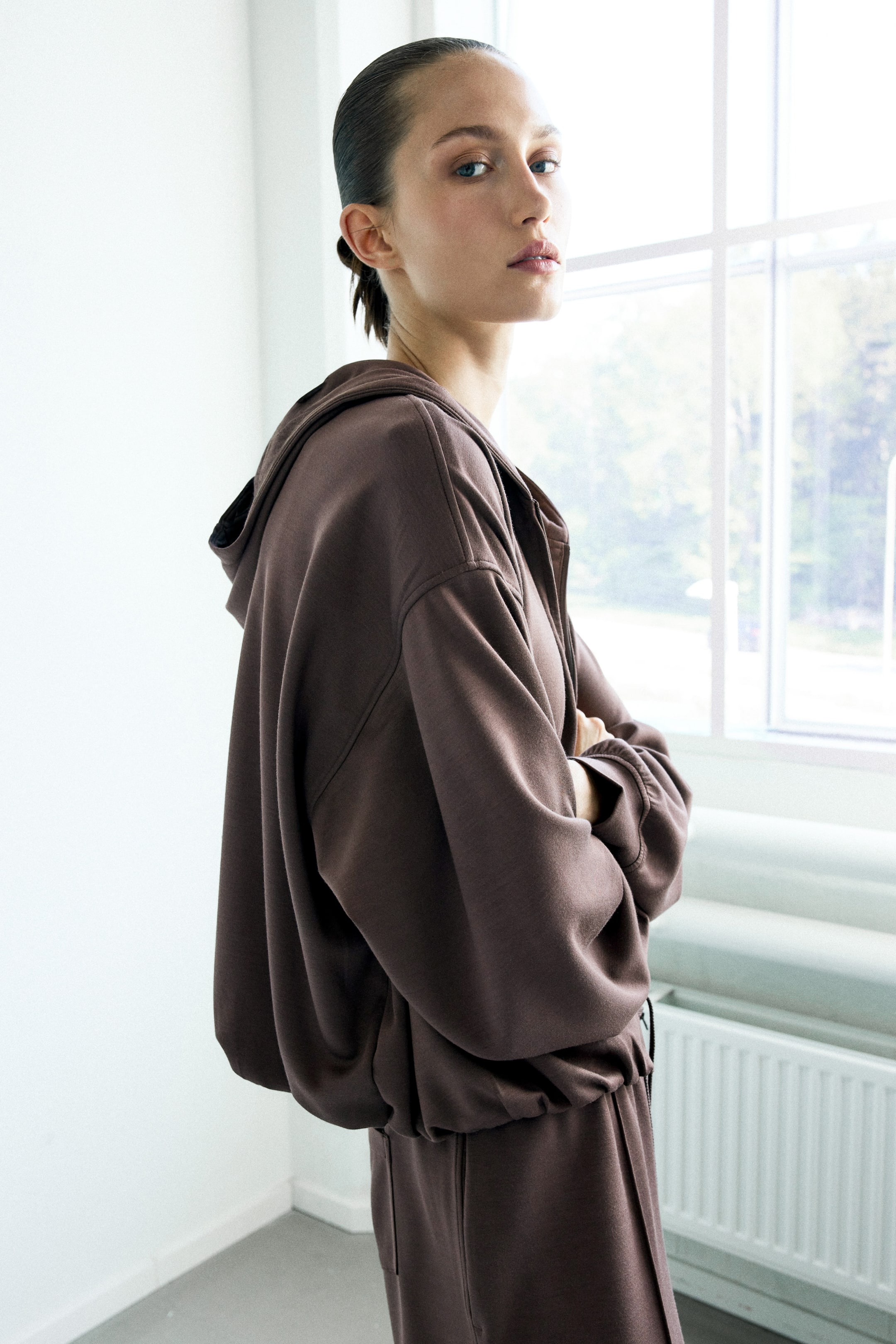 Visa större bild: En brun oversized zip-up hoodie bärs, med en huva, nedhasade axlar och elastiska muddar och fåll, vilket skapar en samlad passform. Den kombineras med matchande löst sittande bruna byxor, med produkten sedd från en lätt sidovinkel.