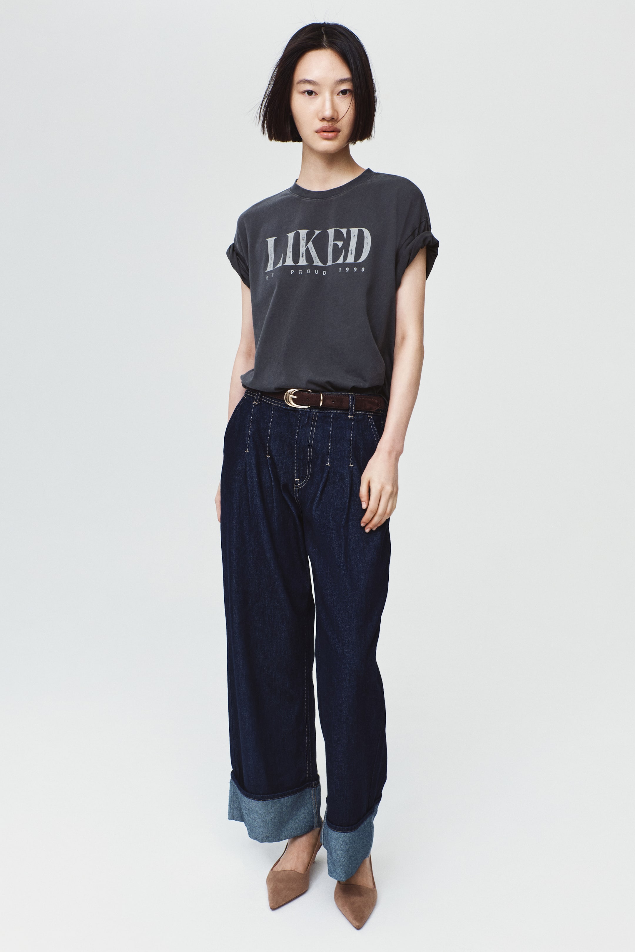 View larger image: The model is wearing an oversized dark gray T-shirt featuring a light-colored printed graphic with the word "LIKED." The T-shirt has a round neckline and is tucked into dark blue wide-leg jeans secured with a brown belt.