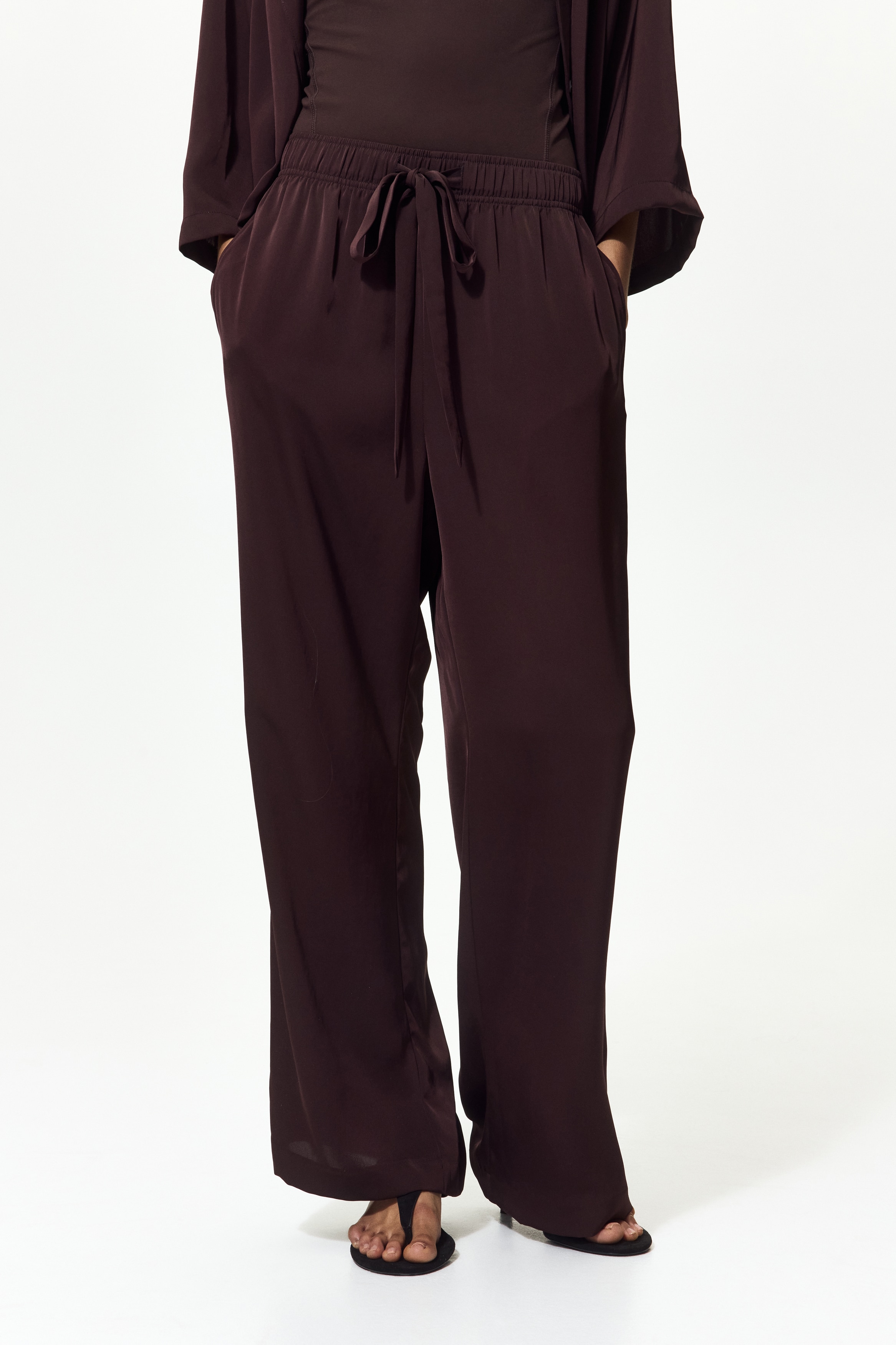 Dark brown wide-leg pants showcase a front view with an elasticized waistband and a matching drawstring tie. The smooth fabric drapes fluidly.