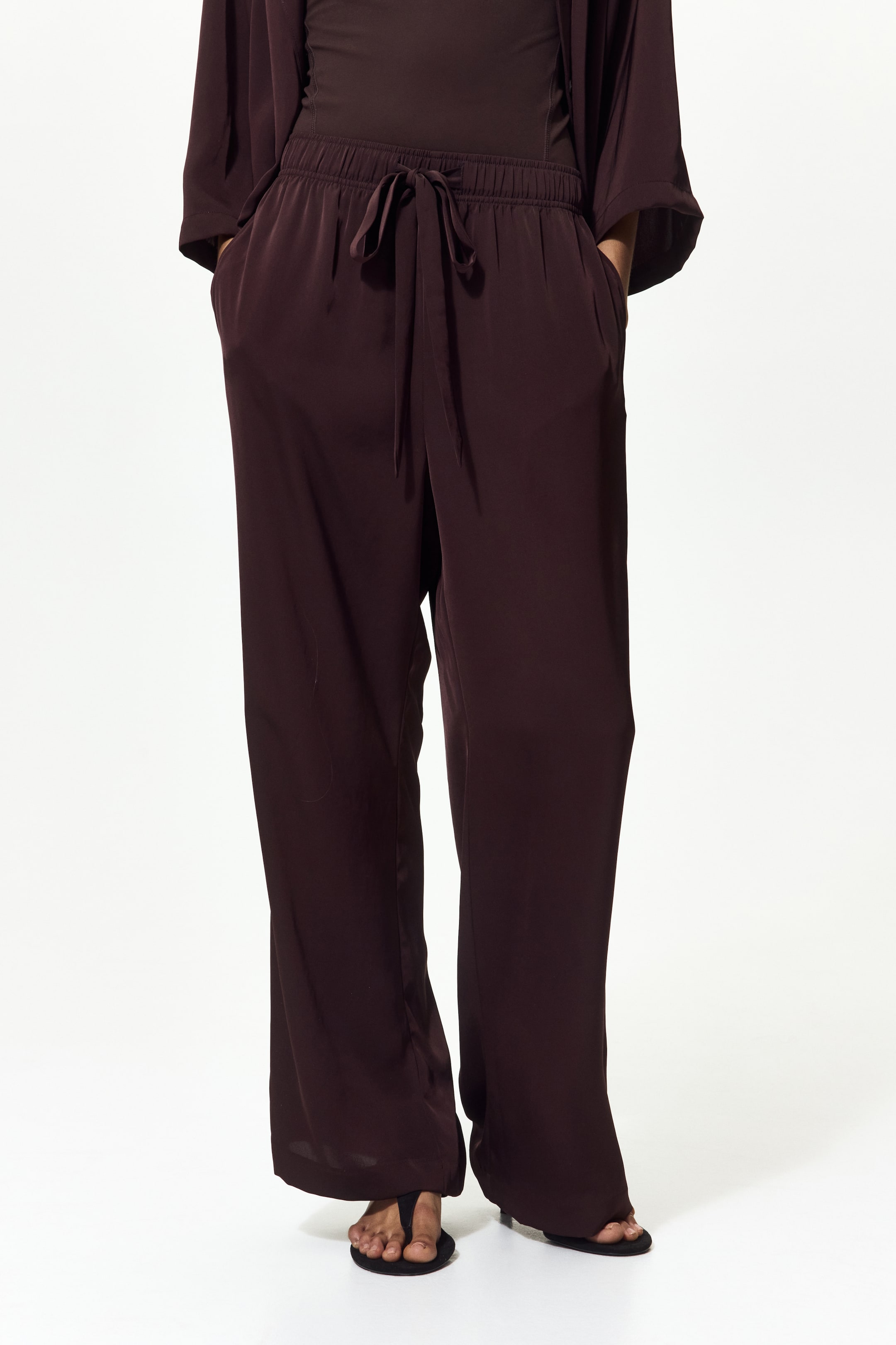 View larger image: The deep brown wide-leg trousers feature an elasticized drawstring waist, with hands comfortably nestled in the side pockets. Their full-length legs elegantly drape over black sandals.