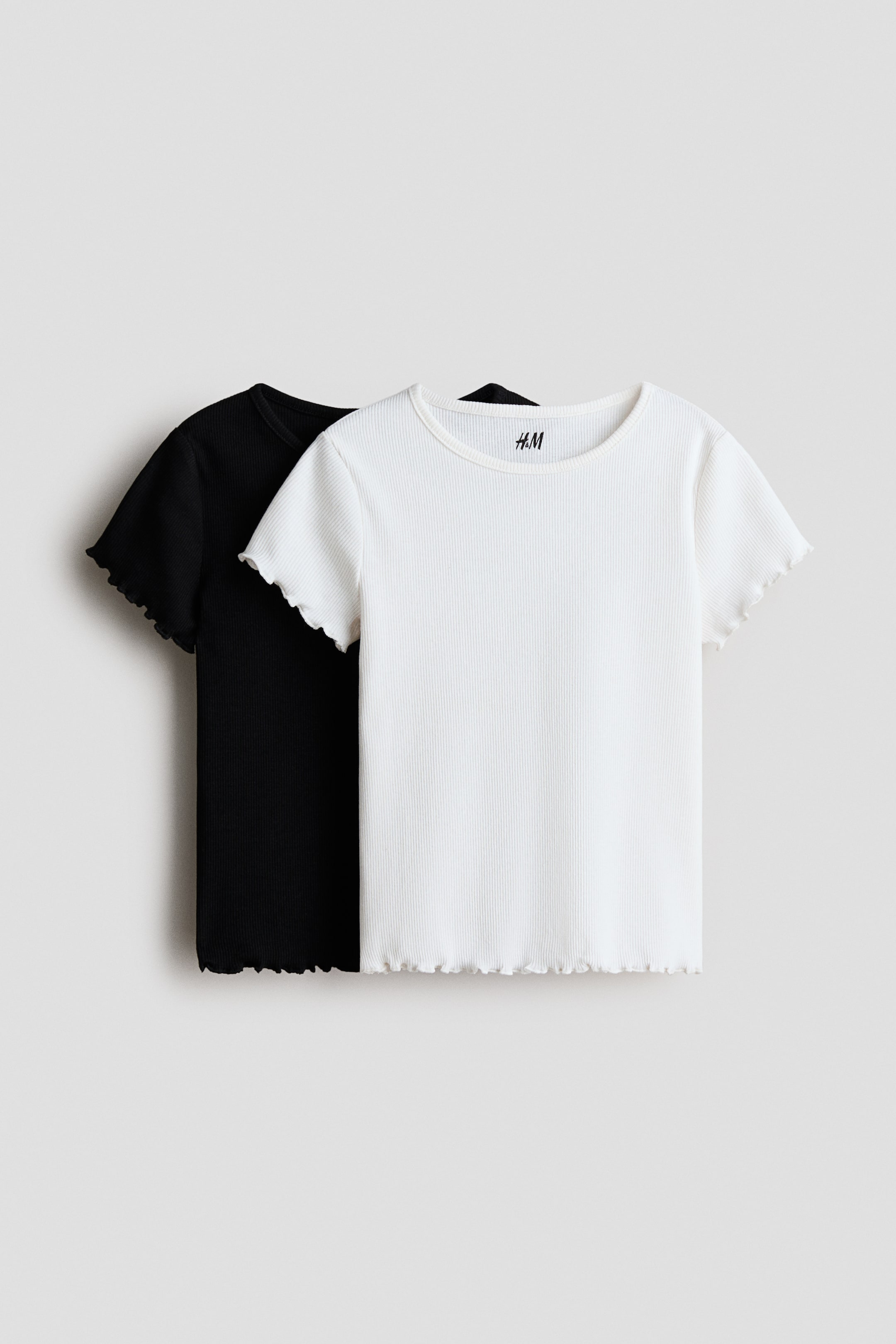 View larger image: Presenting a pair of ribbed short-sleeved t-shirts, showcasing a white t-shirt layered over a black t-shirt. Both t-shirts feature a round neckline and lettuce-edge trim on the short sleeves and bottom hem.