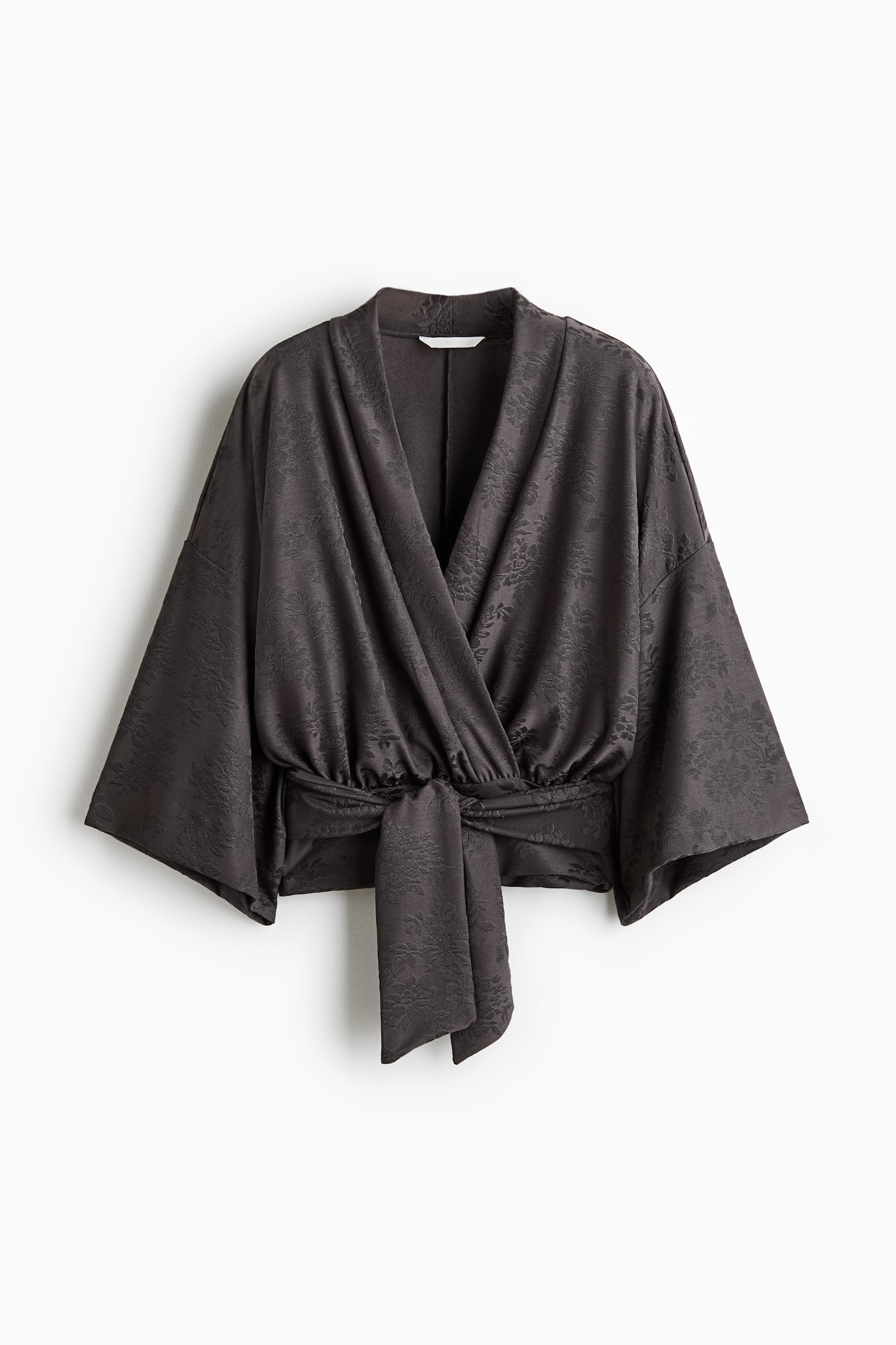 Ciemnoszary kopertowy top jest skierowany przodem, eksponując subtelny kwiatowy wzór żakardowy oraz szerokie rękawy w stylu kimono. Top jest wiązany w talii za pomocą dopasowanego troczka z tego samego materiału, zawiązanego z przodu.