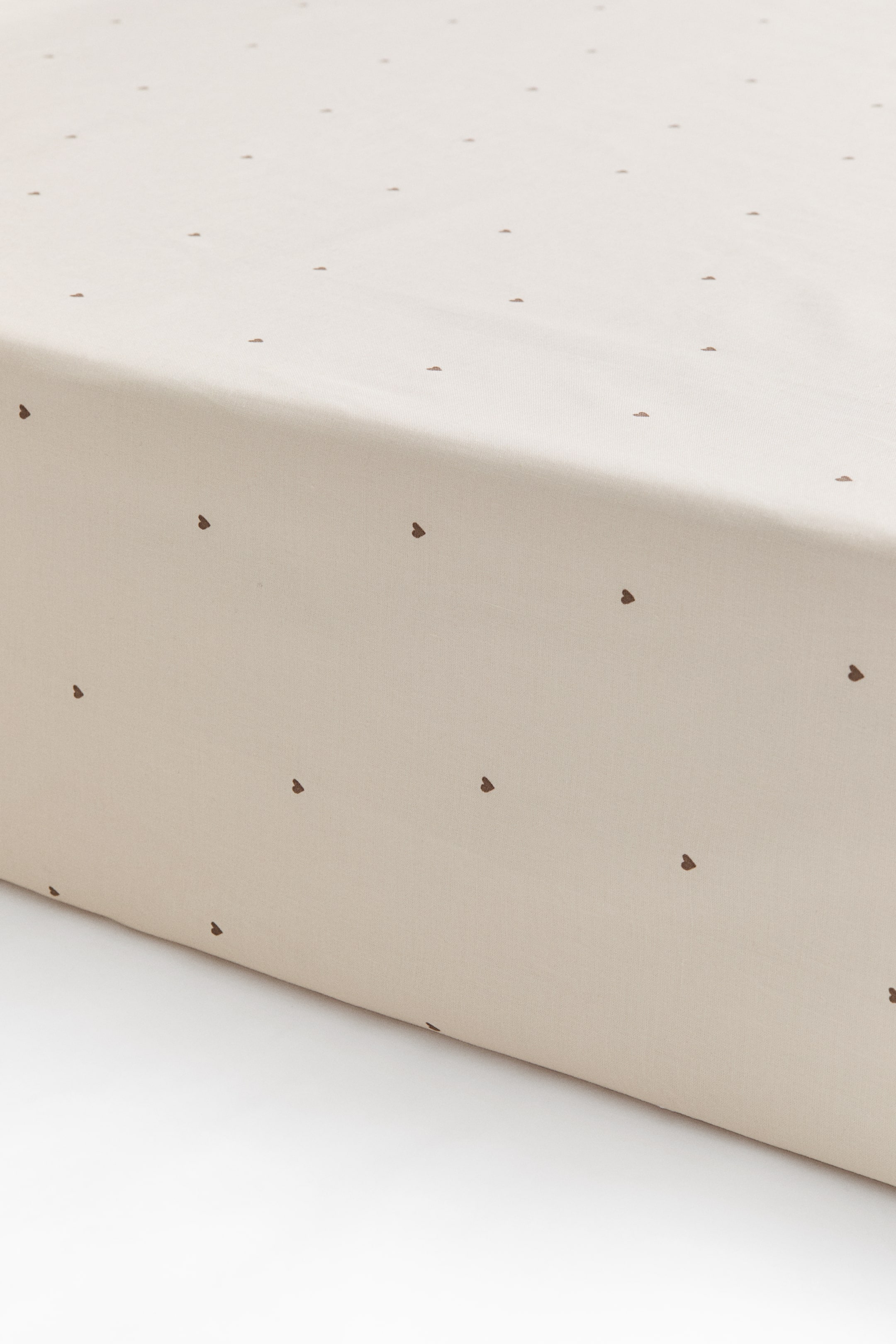 View larger image: A fitted sheet in a light cream cotton weave, featuring a repeating pattern of small dark brown hearts, is covering the corner of a mattress. The close-up view shows the smooth, tailored fit along the top and side edge of the mattress.