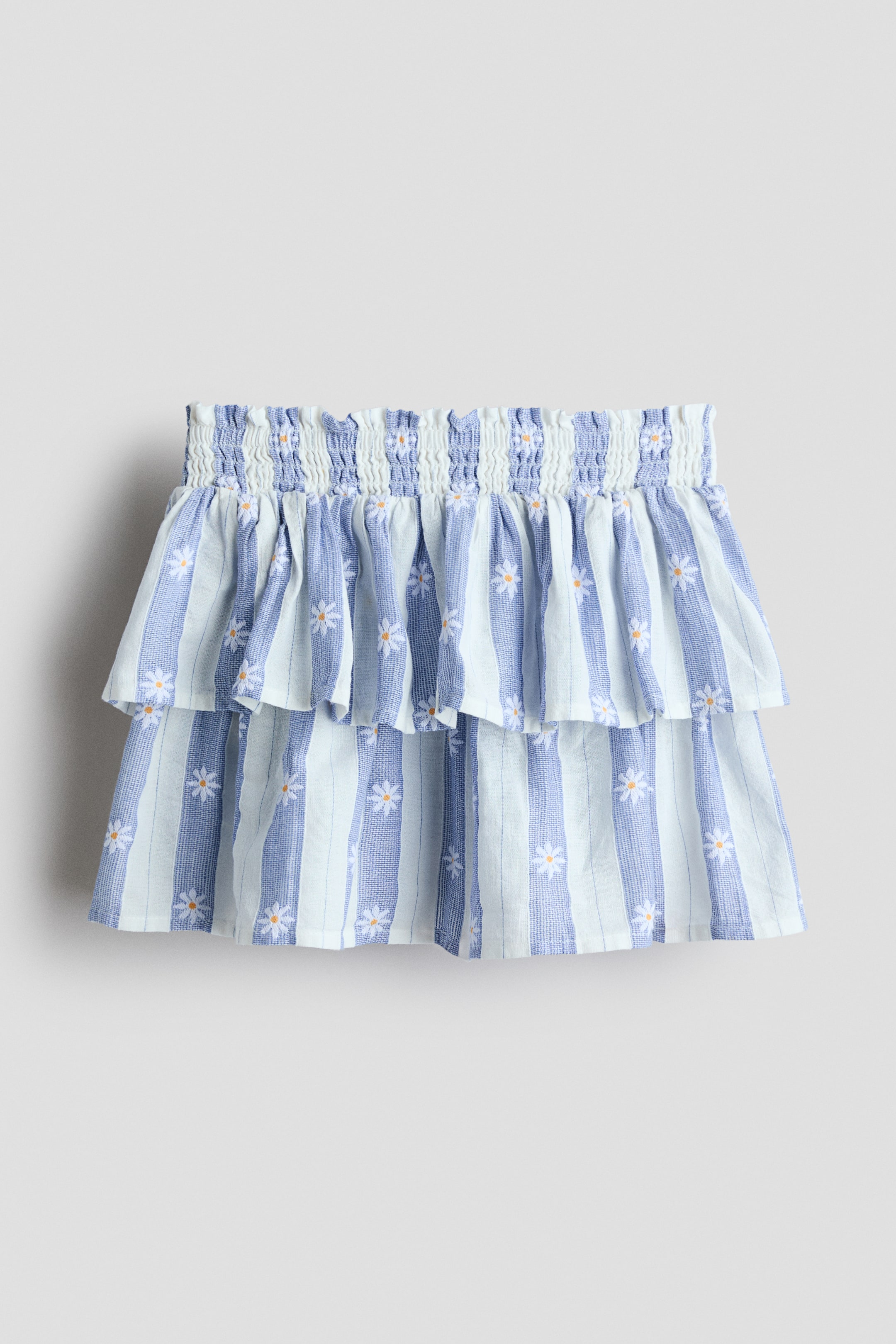 View larger image: The skirt showcases its FRONT, highlighting a shirred elasticated waistband and two ruffled tiers. It flaunts alternating light blue and white vertical stripes, adorned with small white daisy flowers embellishing the fabric.