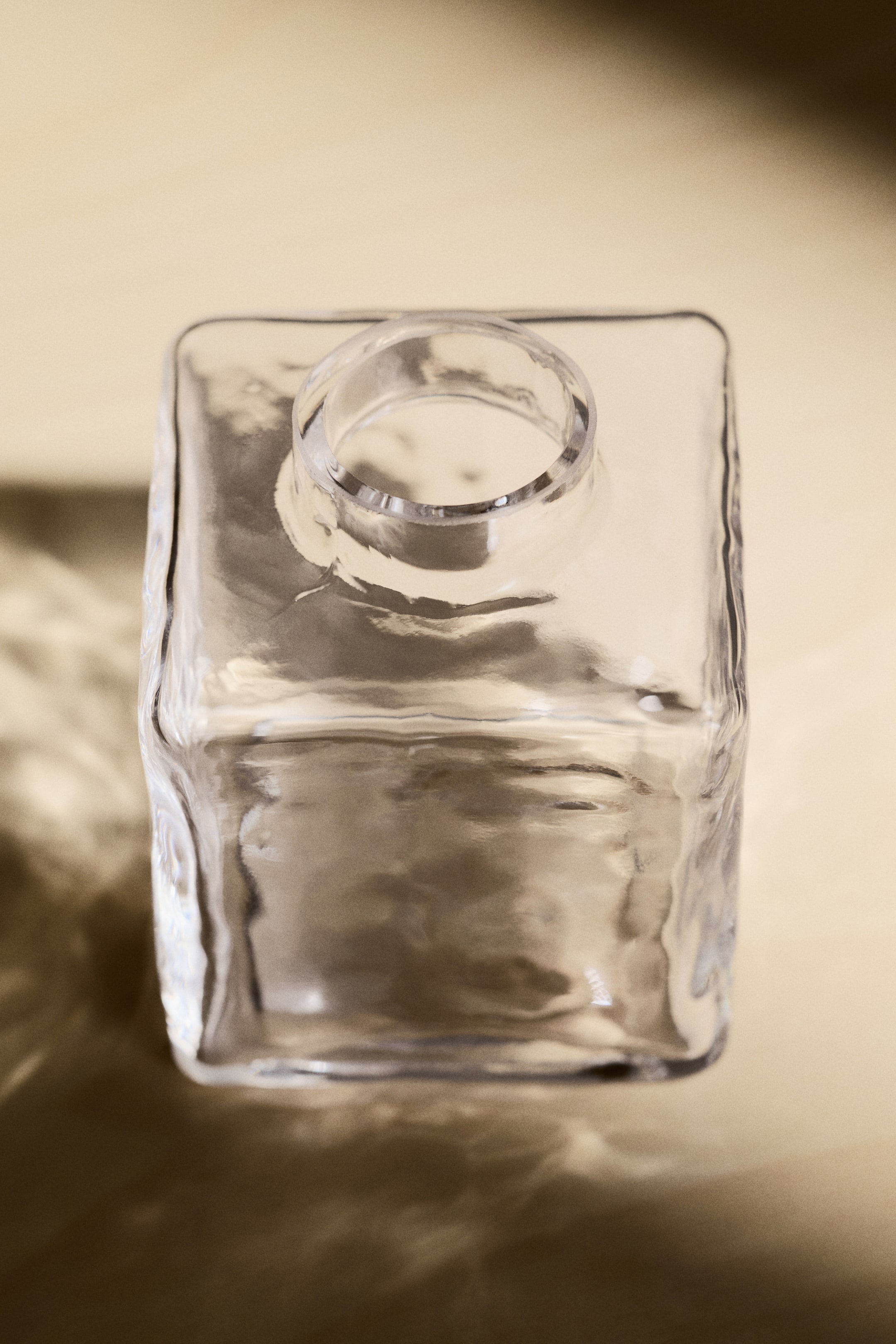 Se større billede: En klar glasflaske ses fra en top-down vinkel, med en firkantet krop og en rund åbning i toppen. Dens gennemsigtige overflade viser subtile bølger og tekstur.