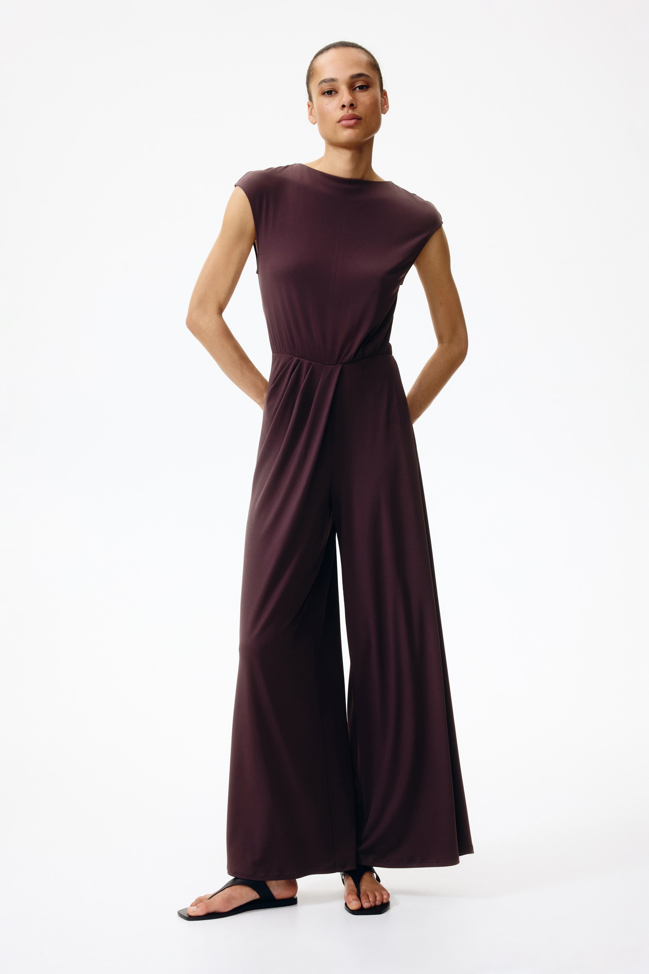 View larger image: A dark plum jumpsuit featuring a boat neckline and cap sleeves, cinching at the waist, and flowing into wide-leg pants, with its full design facing forward.