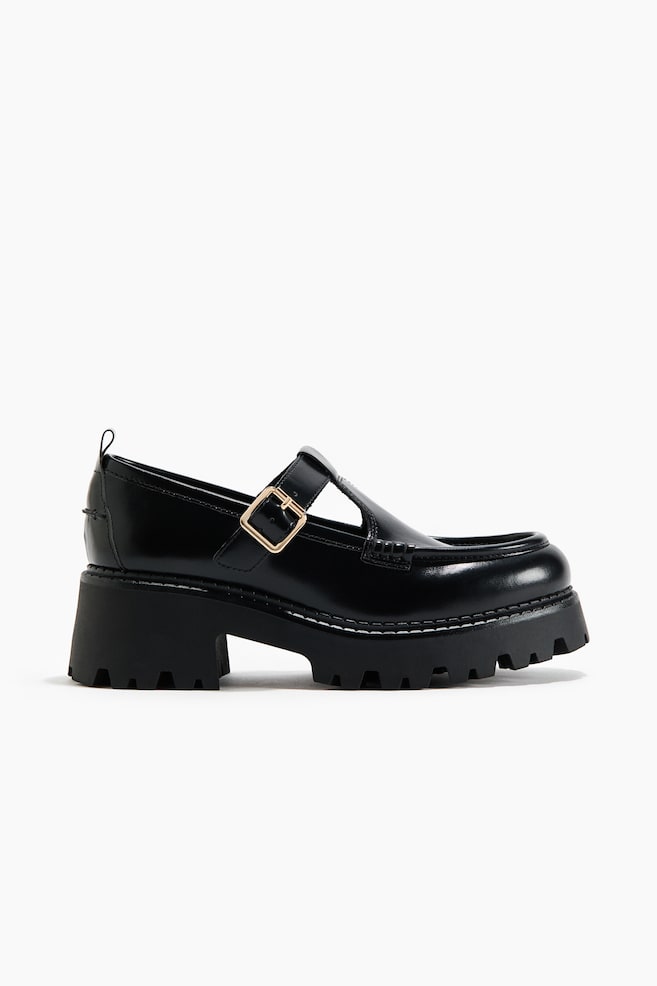 A black Mary Jane shoe is showcasing a glossy finish, viewed from the side. It features a T-strap with a gold-tone buckle and a prominent chunky lug sole with a block heel, highlighted by white stitching at the welt. - 1