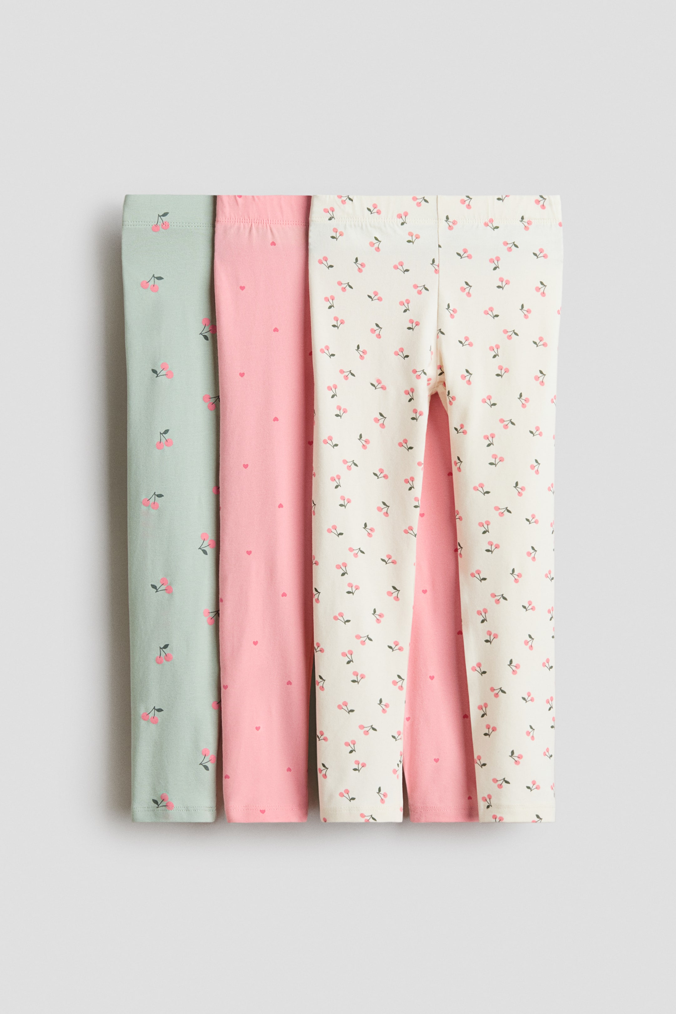 View larger image: The leggings are presented as a set of three, each folded lengthwise. The leftmost pair is light green with pink cherry motifs, the middle pair is pink with small pink hearts, and the rightmost pair is cream with pink cherry motifs. All three pairs feature an elastic waistband.