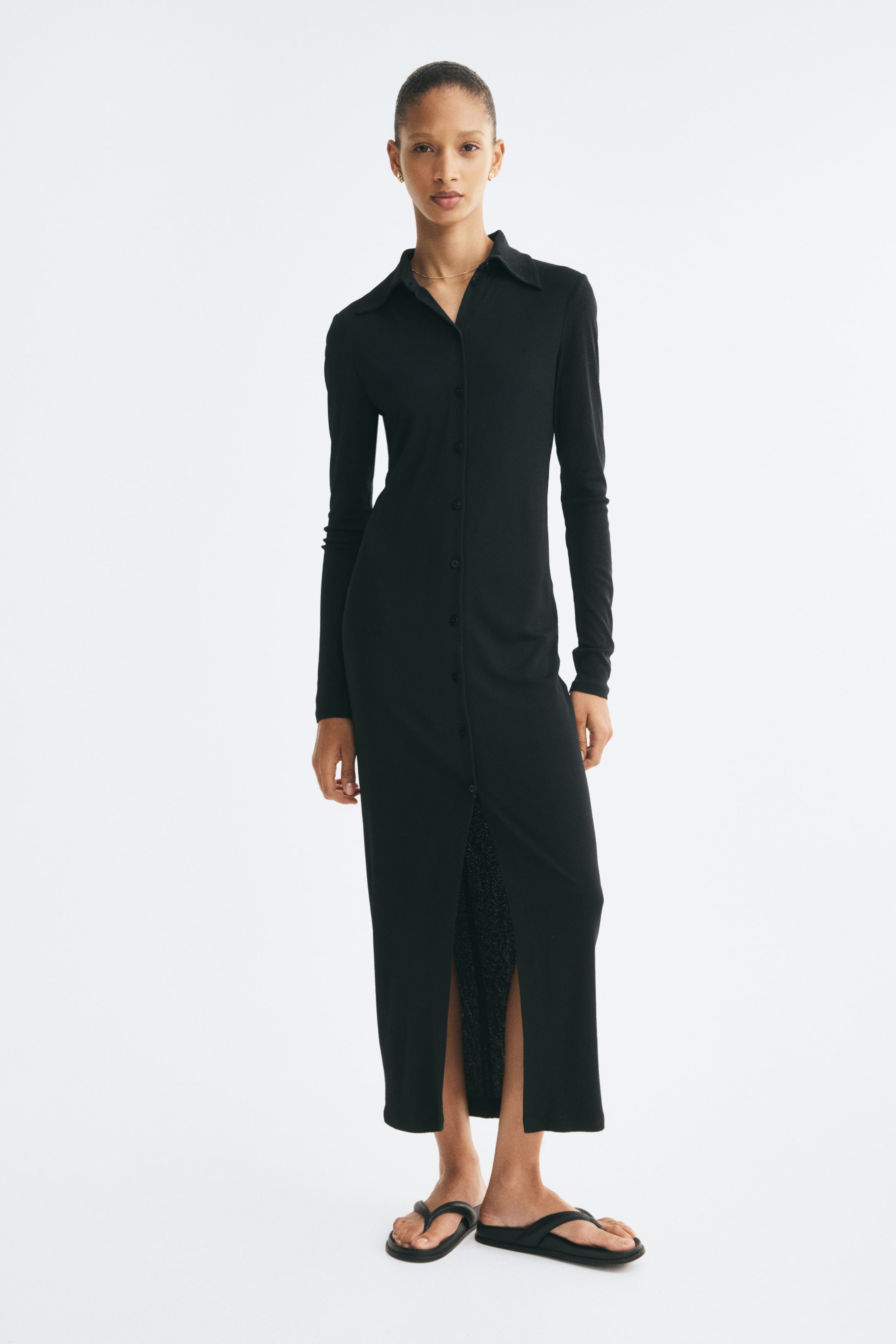 Agrandir l'image: Une longue robe noire ajustée en jersey côtelé est présentée de face, avec un col chemise classique, des manches longues et une fermeture boutonnée sur le devant. La robe présente une silhouette slim qui descend jusqu’aux chevilles, avec une fente sur le devant créée par la partie inférieure laissée déboutonnée.