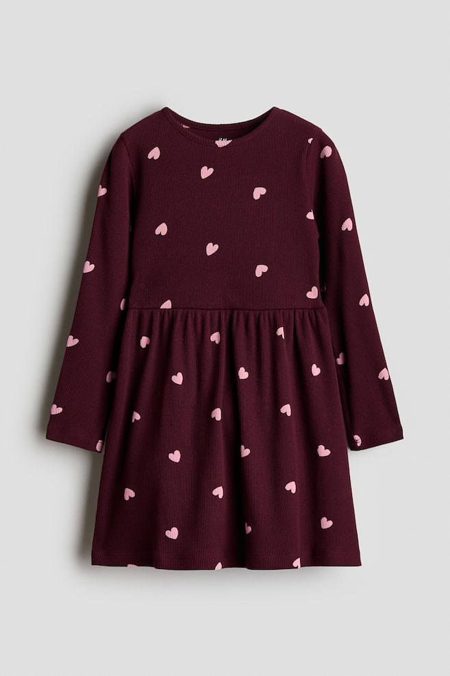 En mÃ¶rk vinrÃ¶d ribbstickad klÃ¤nning med ett heltÃ¤ckande mÃ¶nster av smÃ¥ rosa hjÃ¤rtan hÃ¤nger vÃ¤nd mot framsidan. KlÃ¤nningen visar upp en rund halsringning, lÃ¥nga Ã¤rmar, en rynkad sÃ¶m i midjan och en lÃ¤tt utsvÃ¤ngd kjol. - 1