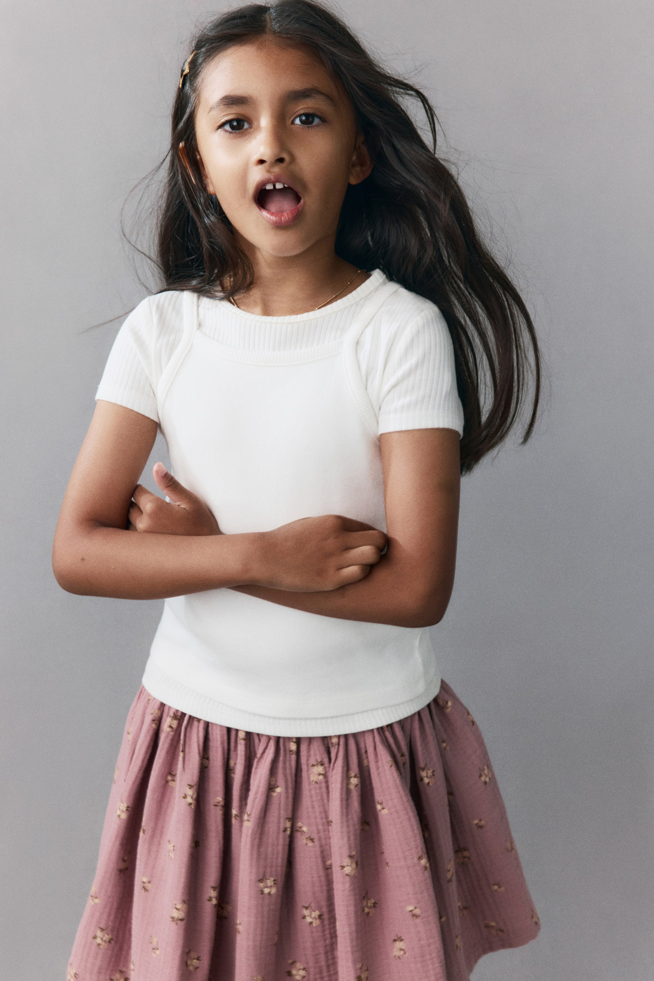 View larger image: A flared skirt in dusty pink cotton muslin with an all-over floral print is displayed from the front. The skirt features a gathered waist that adds volume and is styled with a white ribbed T-shirt.
