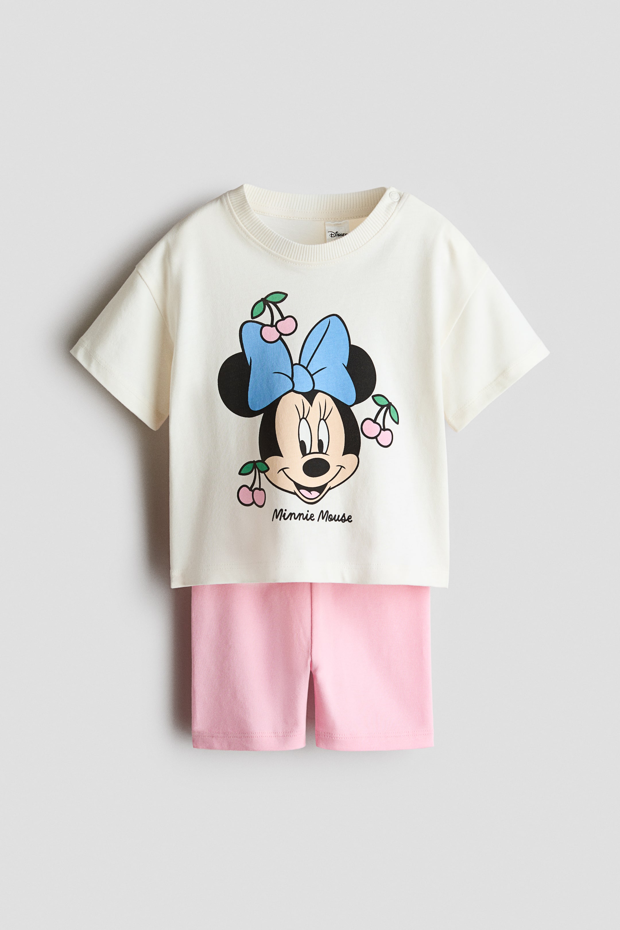 View larger image: A 2-piece cotton set features an off-white short-sleeved t-shirt and pink shorts, both facing forward. The t-shirt showcases a Minnie Mouse graphic with a blue bow and pink cherries, and displays a ribbed crew neck with snaps on the left shoulder.