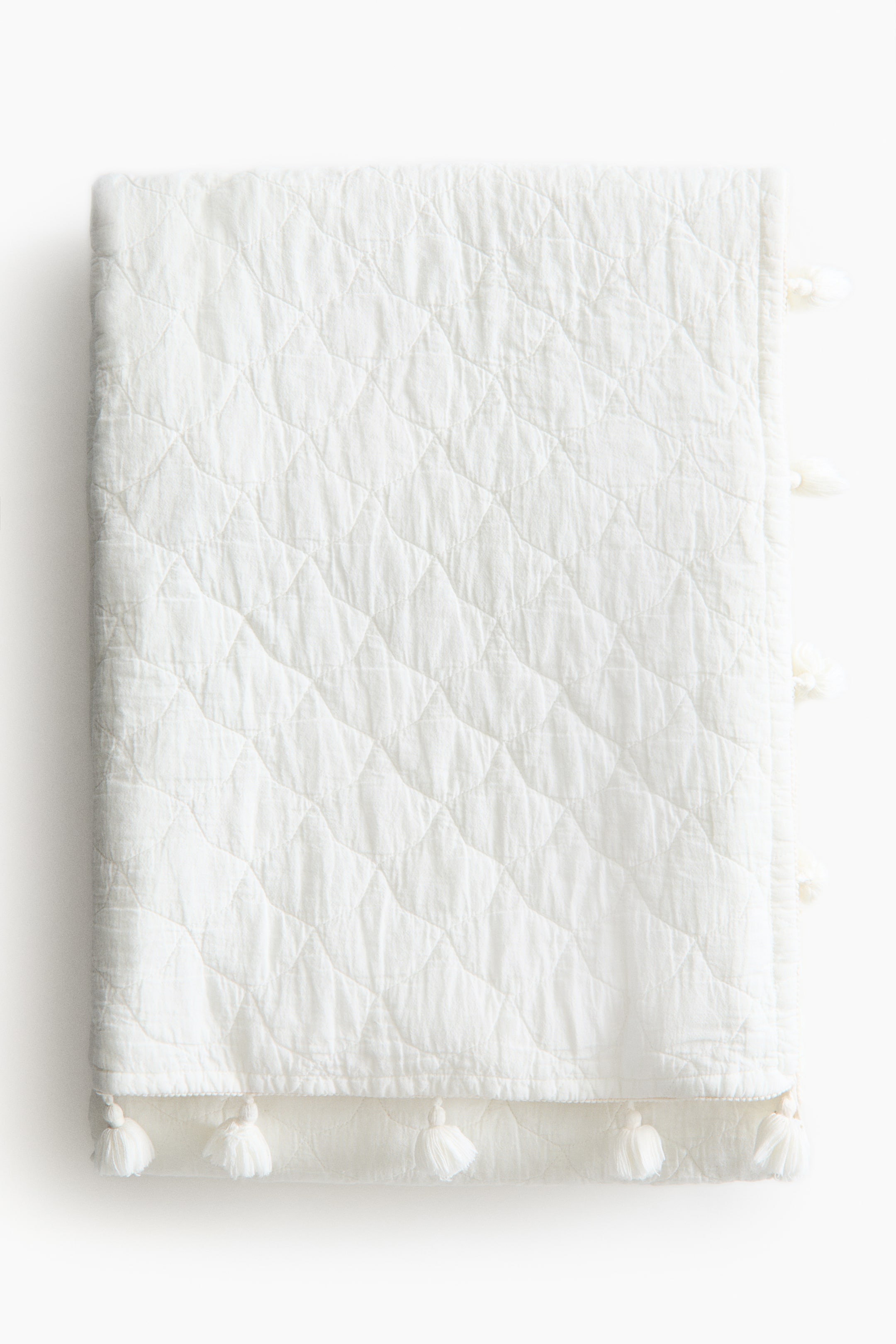 Zobacz większy obraz: Lekko pikowane narzuty na łóżko są prezentowane przodem, z pikowaną bawełnianą tkaniną o subtelnym, falistym wzorze. Z przodu ich widocznych boków i dolnej krawędzi zwisają chwosty.