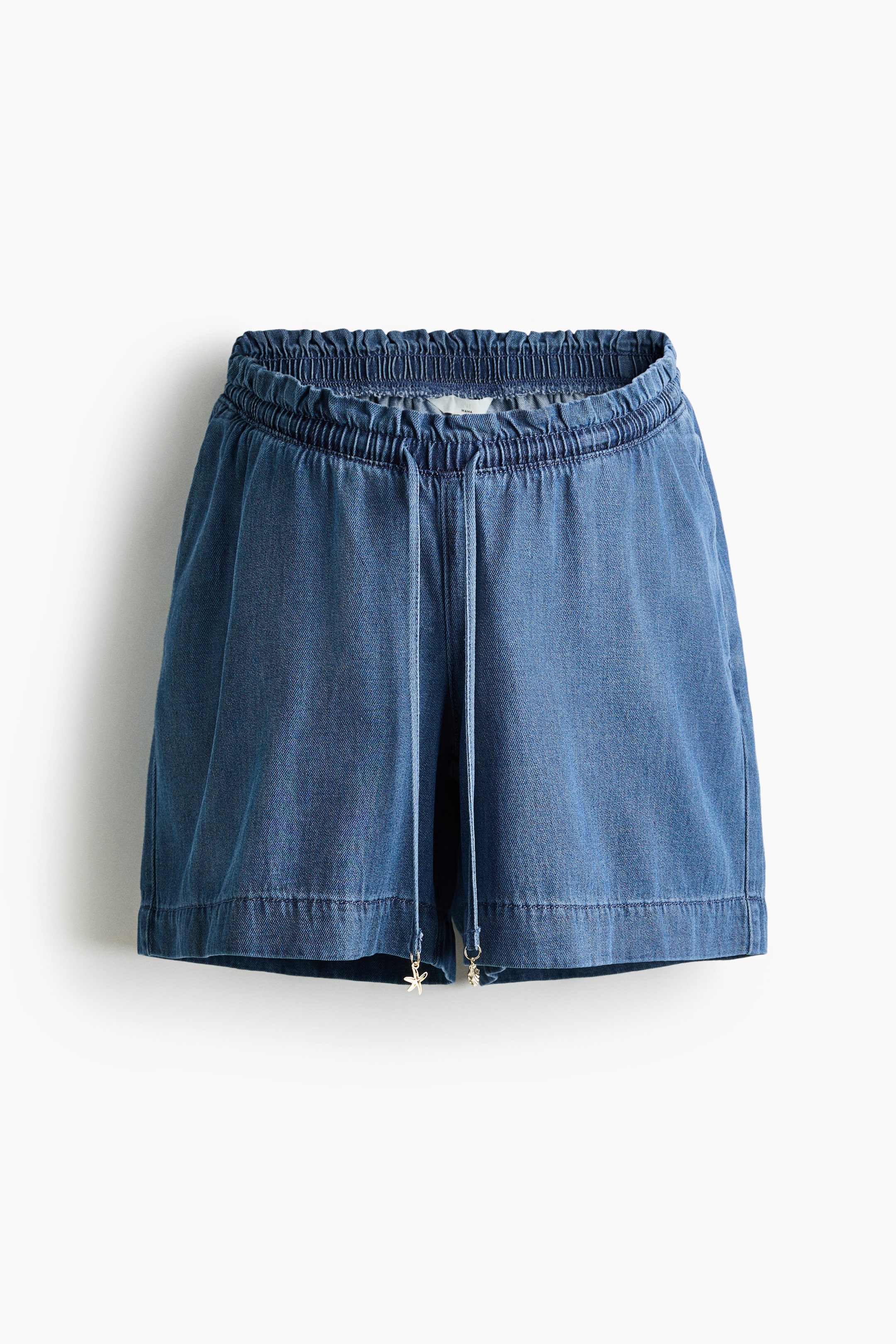 View larger image: Blue denim shorts feature an elasticized, shirred waistband with a front drawstring. Small metallic charms adorn the drawstring ends, and the shorts finish with folded hems.