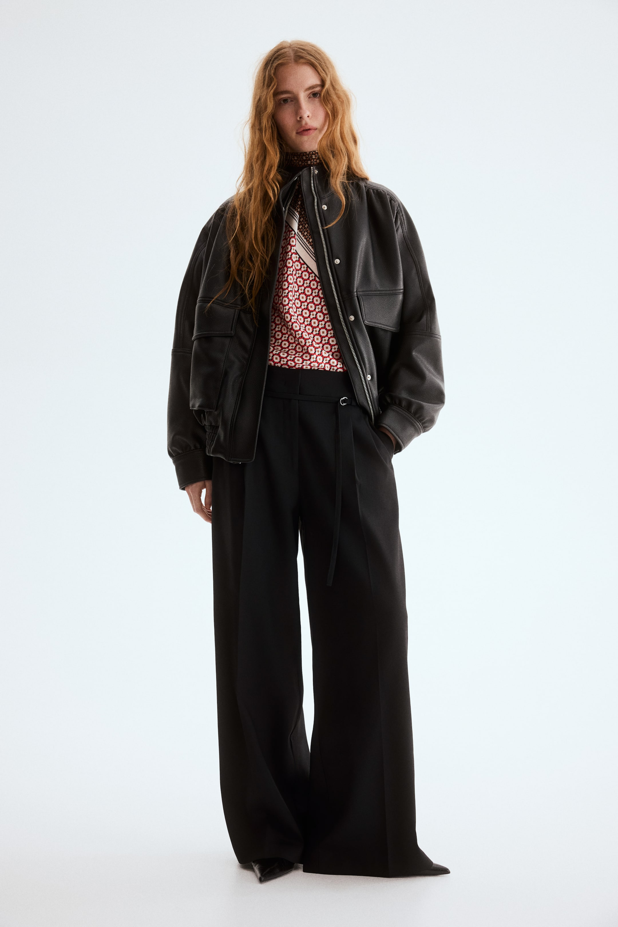 查看大圖: 黑色、寬鬆剪裁的斜紋布長褲由模特穿著，特點是高腰前褶、寬腿前摺，以及配有D形環的窄腰帶。模特的右手放在斜側口袋中。