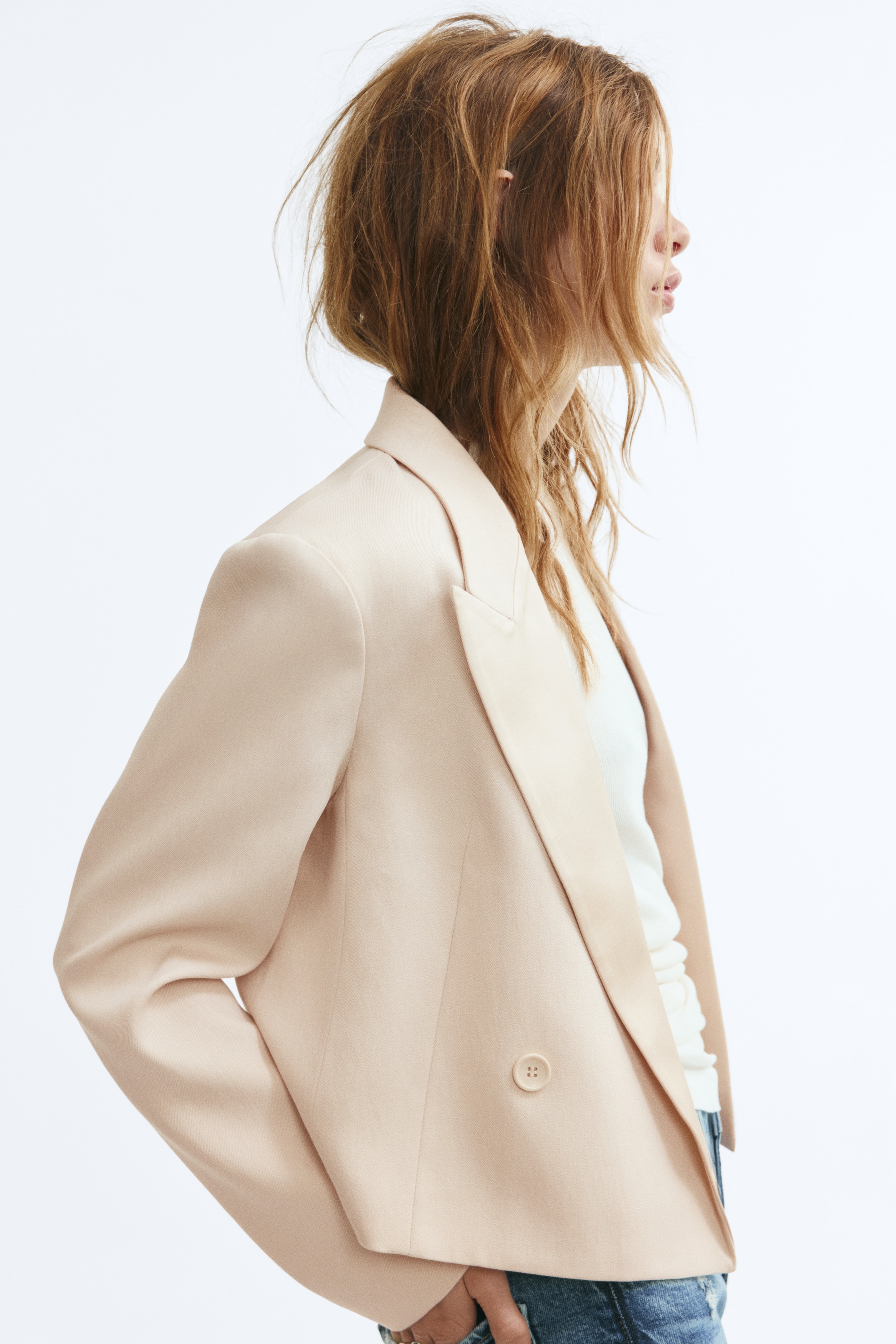 A light beige cropped blazer is facing front, showcasing its structured silhouette with wide peak lapels and padded shoulders. The design features long sleeves, a double-breasted front with two tonal buttons, and clean-cut angled seam details.