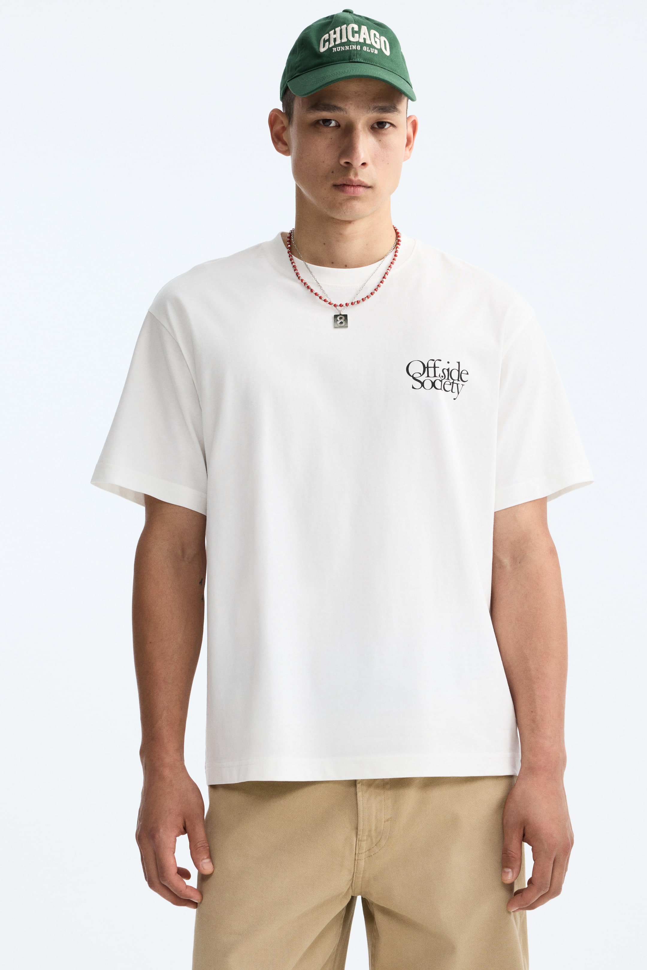 View larger image: A relaxed-fit t-shirt in off-white, facing forward, featuring a crew neck and short sleeves, with the black "Offside Society" script text on the left chest. A red beaded necklace drapes over the t-shirt, extending over khaki-colored pants.