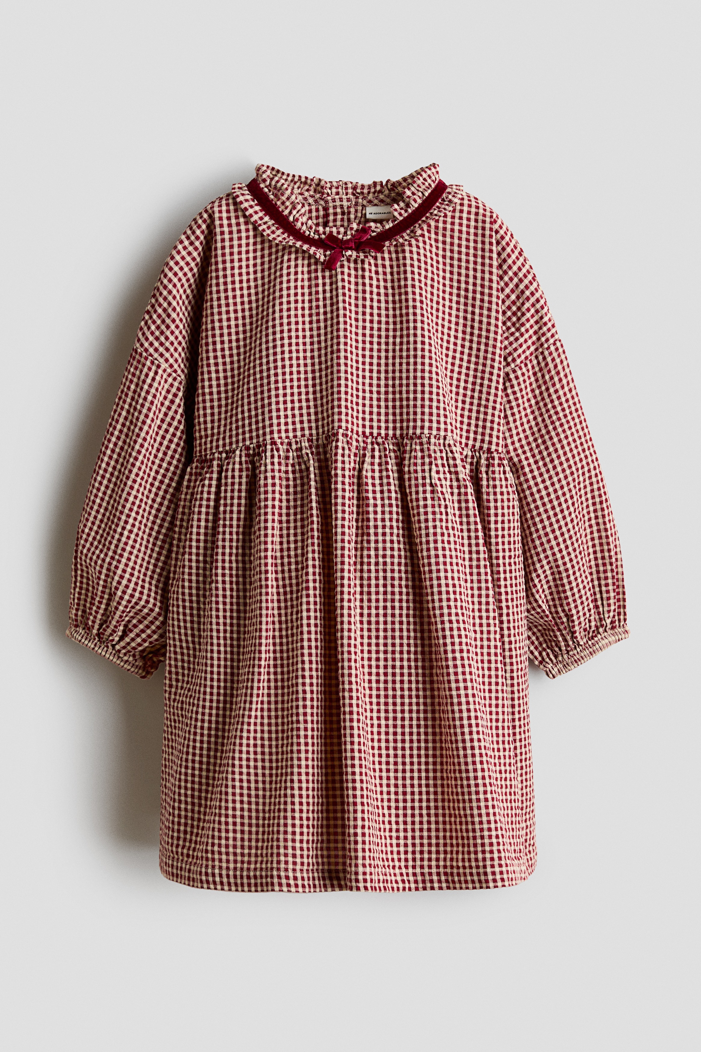 KÄ±rmÄ±zÄ± ve beyaz gingham seersucker bir elbise, bol ve kloÅ siluetini Ã¶ne Ã§Ä±karmak iÃ§in Ã¼Ã§ Ã§eyrek yan aÃ§Ä±dan gÃ¶steriliyor. Elbise; koyu kÄ±rmÄ±zÄ± kadife biye ve kÃ¼Ã§Ã¼k fiyonk detaylÄ± fÄ±rfÄ±rlÄ± yaka, dar lastikli manÅetlere sahip uzun ve hacimli kollar ve bel kÄ±smÄ±nda bÃ¼zgÃ¼ler ile tasarlanmÄ±ÅtÄ±r.