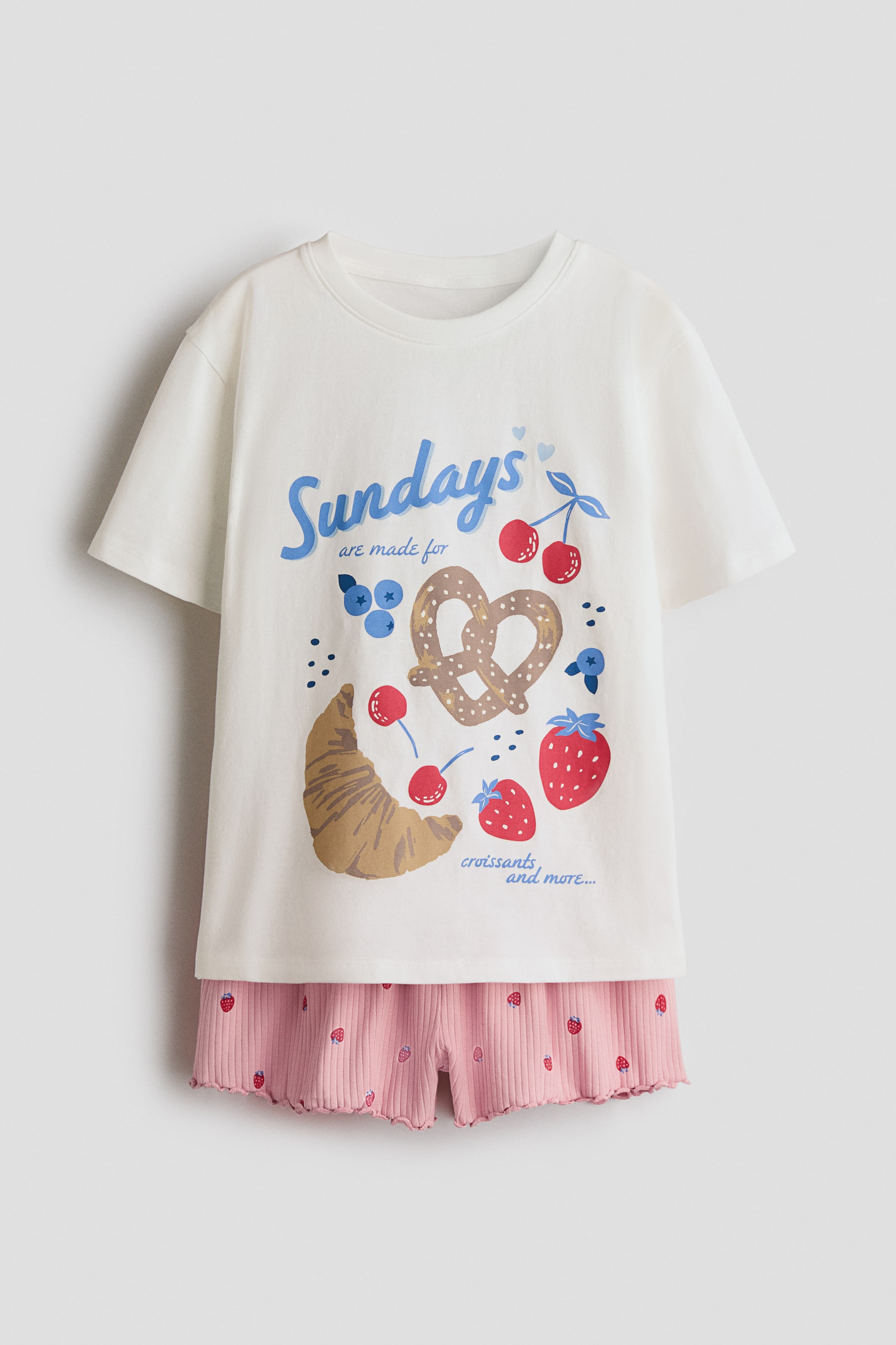 Visualizza l'immagine ingrandita: I pigiami presentano una T-shirt color bianco sporco e pantaloncini rosa, presentati dal davanti. La T-shirt mostra uno scollo rotondo, un orlo dritto e una grande stampa grafica frontale con la scritta "Le domeniche sono fatte per i croissant e altro ancora..." accompagnata da illustrazioni di un croissant, un pretzel, ciliegie, fragole e mirtilli. I pantaloncini rosa presentano una texture a coste con piccole stampe di fragole, una vita elasticizzata e un orlo smerlato.