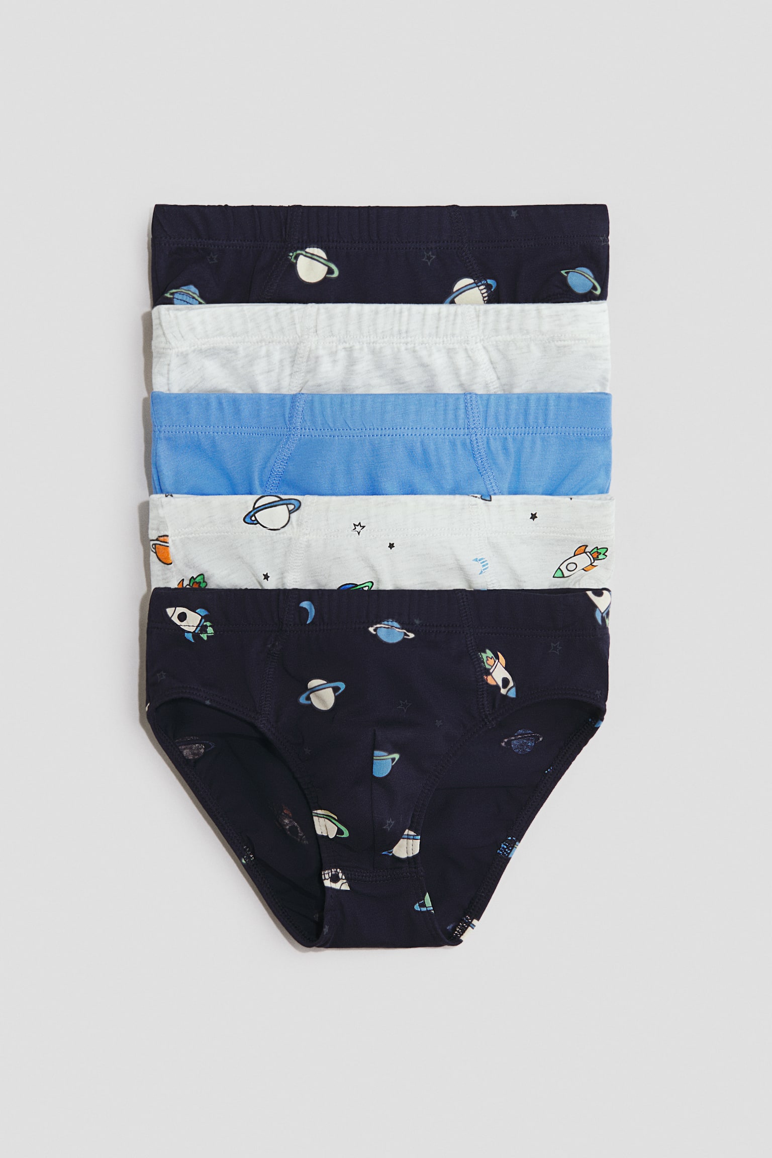 A five-pack of boys' briefs is displayed in a stacked, front-facing arrangement. The set includes solid light grey and blue pairs, as well as navy and white pairs decorated with an all-over space-themed print featuring planets, rockets, and stars.
