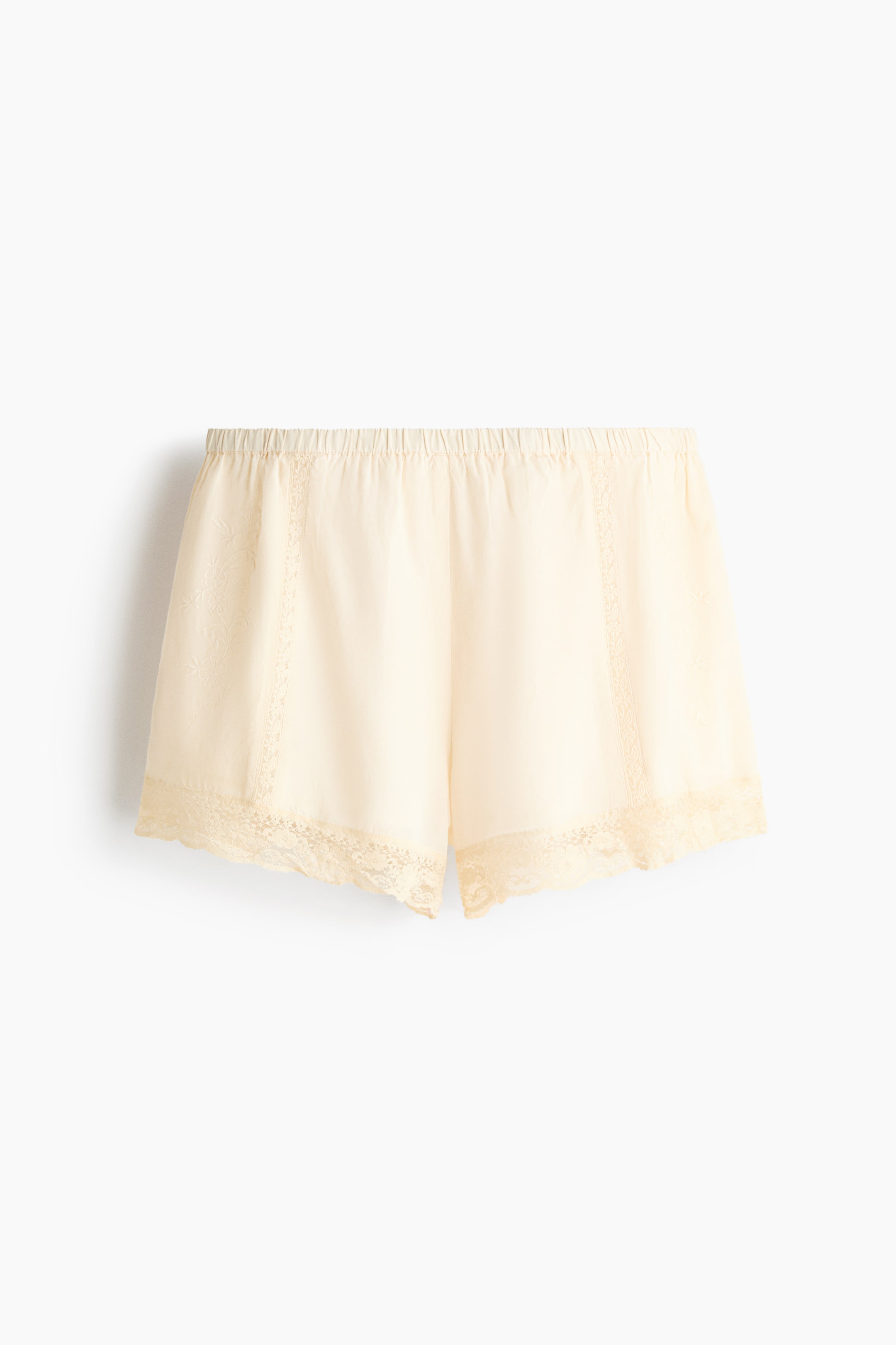 Visualizza immagine più grande: Shorts crema rivolti in avanti, con fascia elastica in vita, bordo in pizzo lungo le aperture delle gambe, pannelli verticali in pizzo sui lati e un delicato motivo floreale sul tessuto.