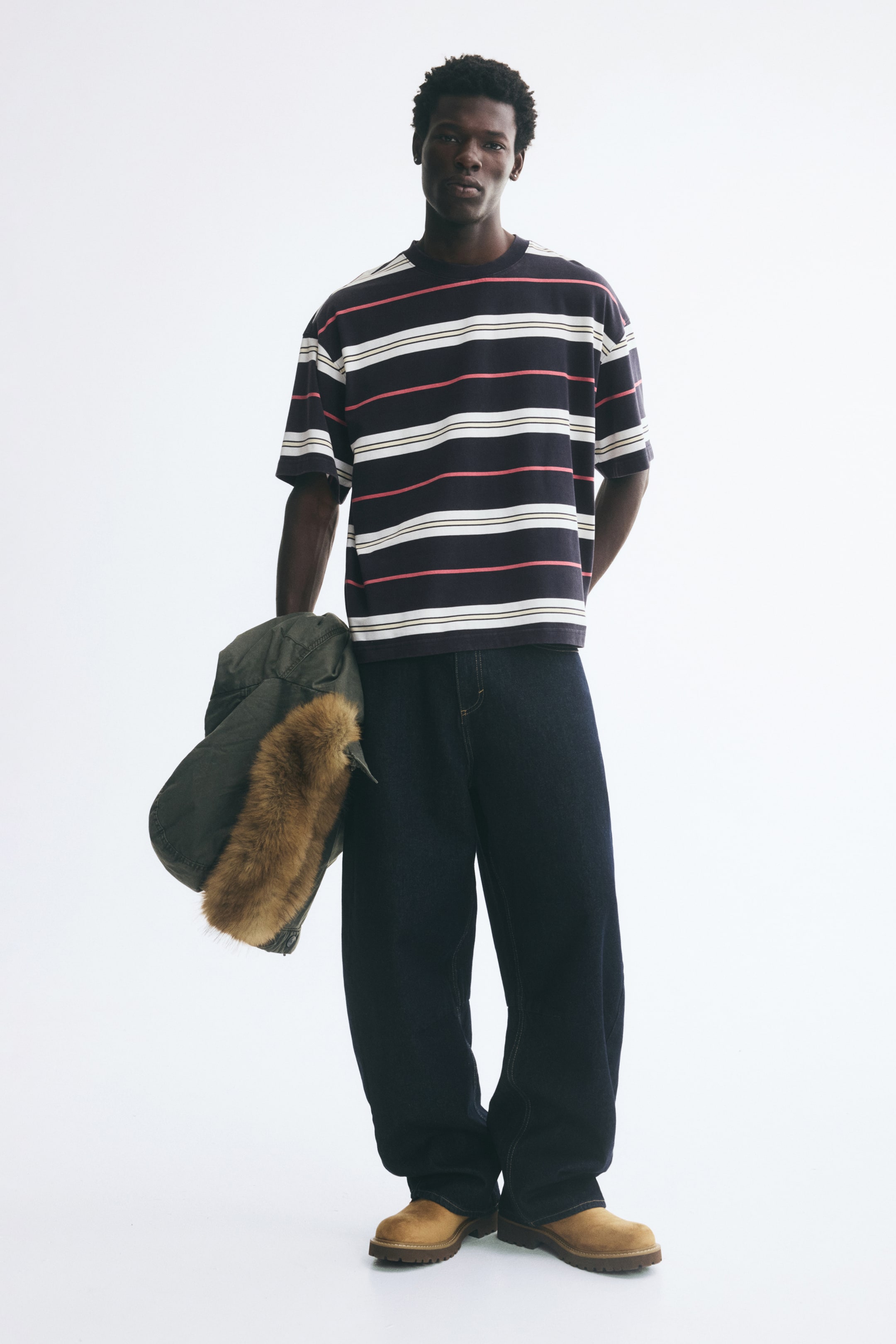 View larger image: An oversized, striped tee in heavyweight cotton jersey is worn by the model, featuring horizontal white, pink, and beige stripes on a dark navy base, with dropped shoulders and a rib-trimmed neckline. The model is styling the baggy tee with loose, dark blue jeans while holding a green jacket with a faux-fur hood.