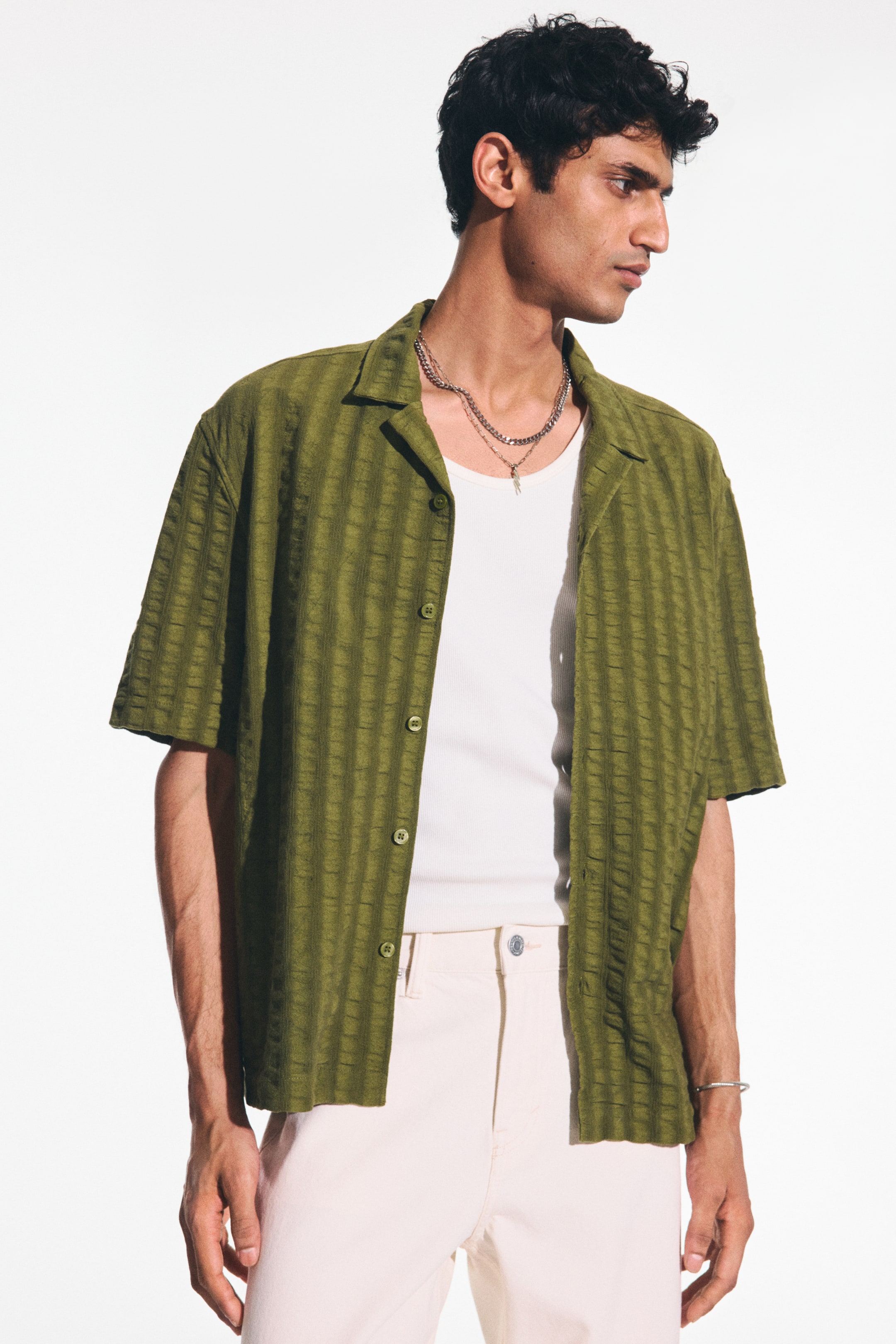 View larger image: An olive green short-sleeved shirt showcases a camp collar and a textured vertical stripe pattern. It offers a relaxed fit with a front button closure, styled open over a white ribbed tank top.
