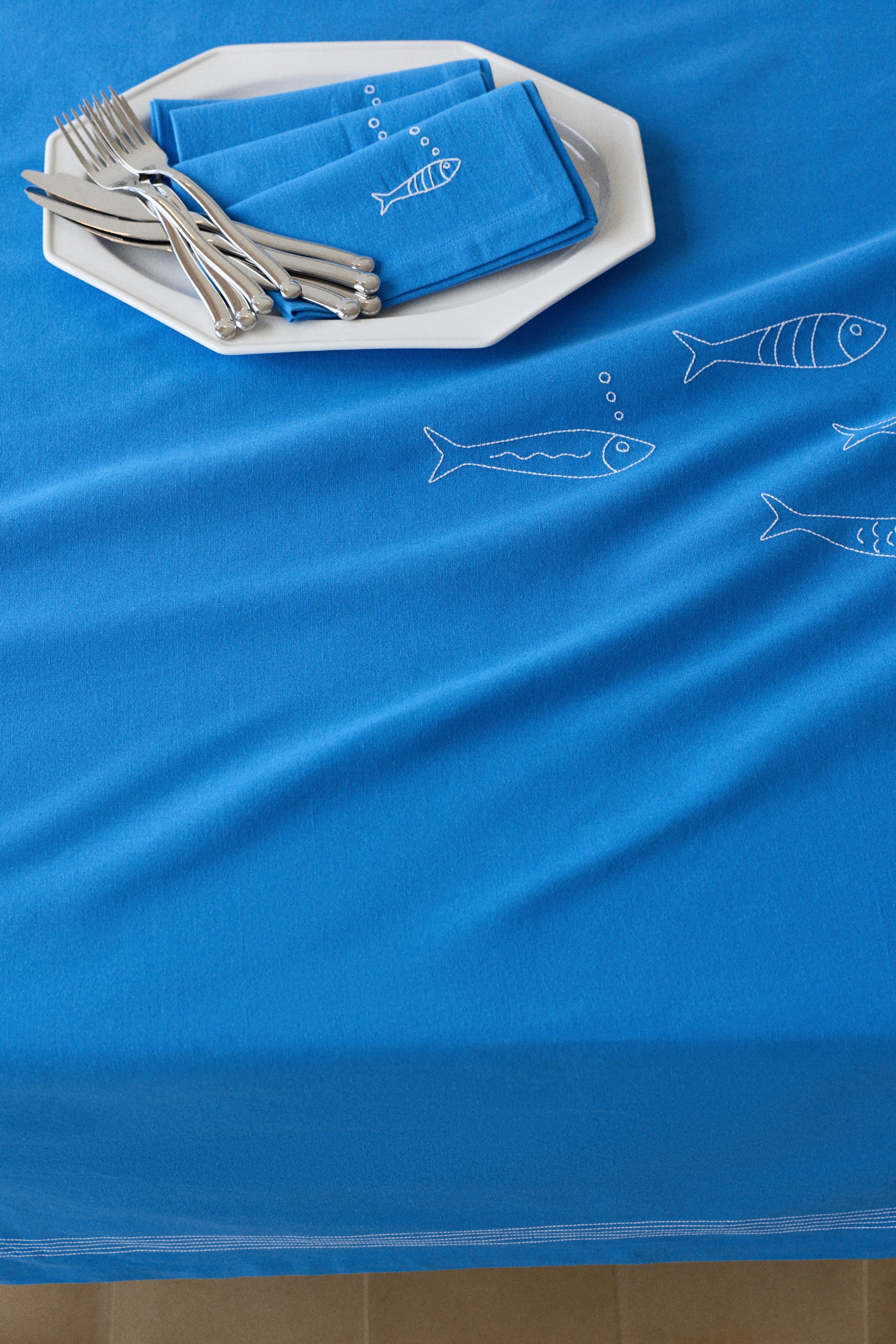 Voir une image plus grande: Une nappe bleue, ornée de contours de poissons blancs brodés avec quelques rayures internes et une bordure doublement cousue, recouvre une surface. Des serviettes bleues assorties, affichant des poissons brodés blancs et des bulles, sont pliées et reposent sur une assiette octogonale blanche avec des couverts.