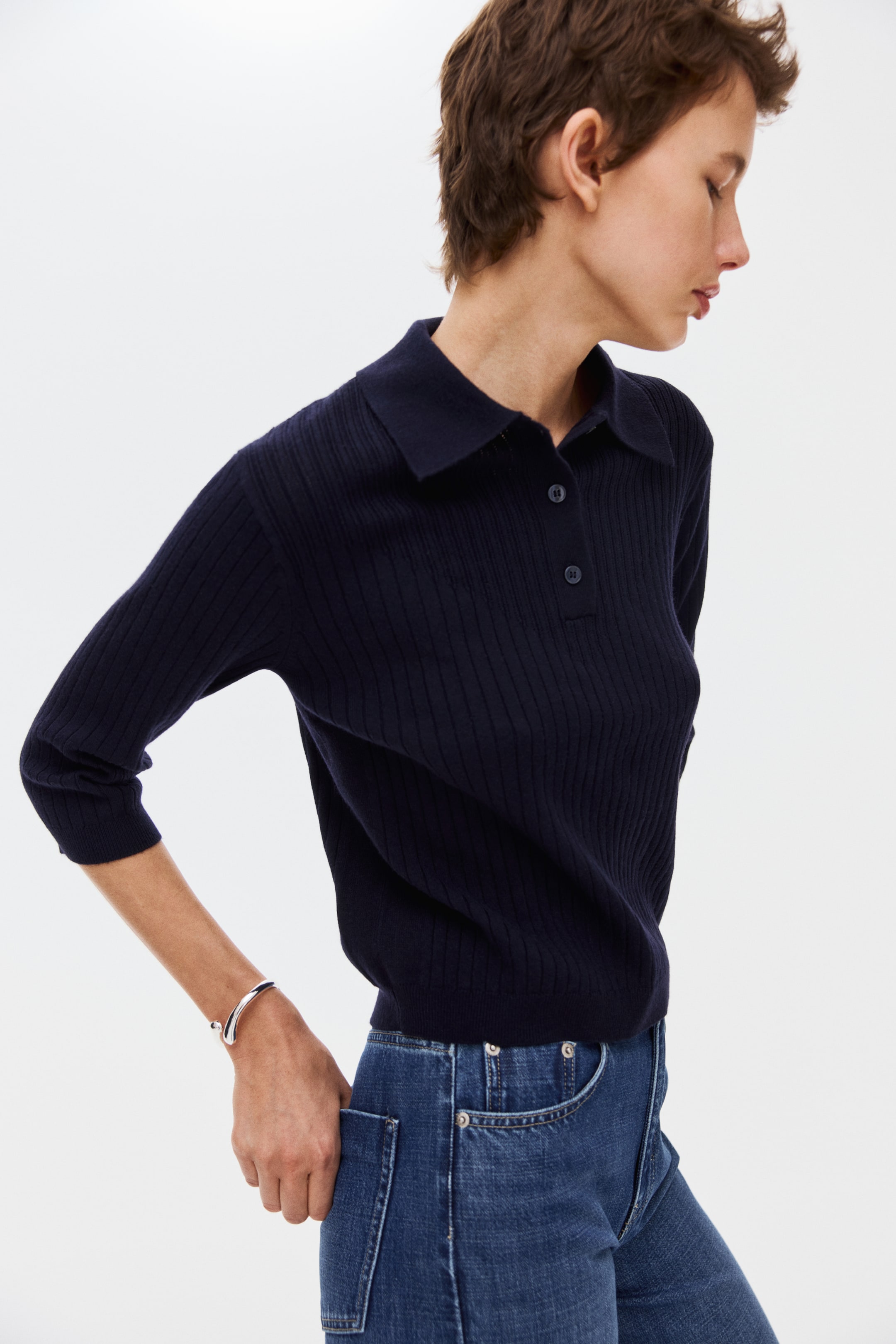 Ver imagem maior: Está a usar um polo de malha canelada azul-marinho, com gola e pala com três botões. Tem mangas três-quartos e a bainha está enfiada em jeans azuis.