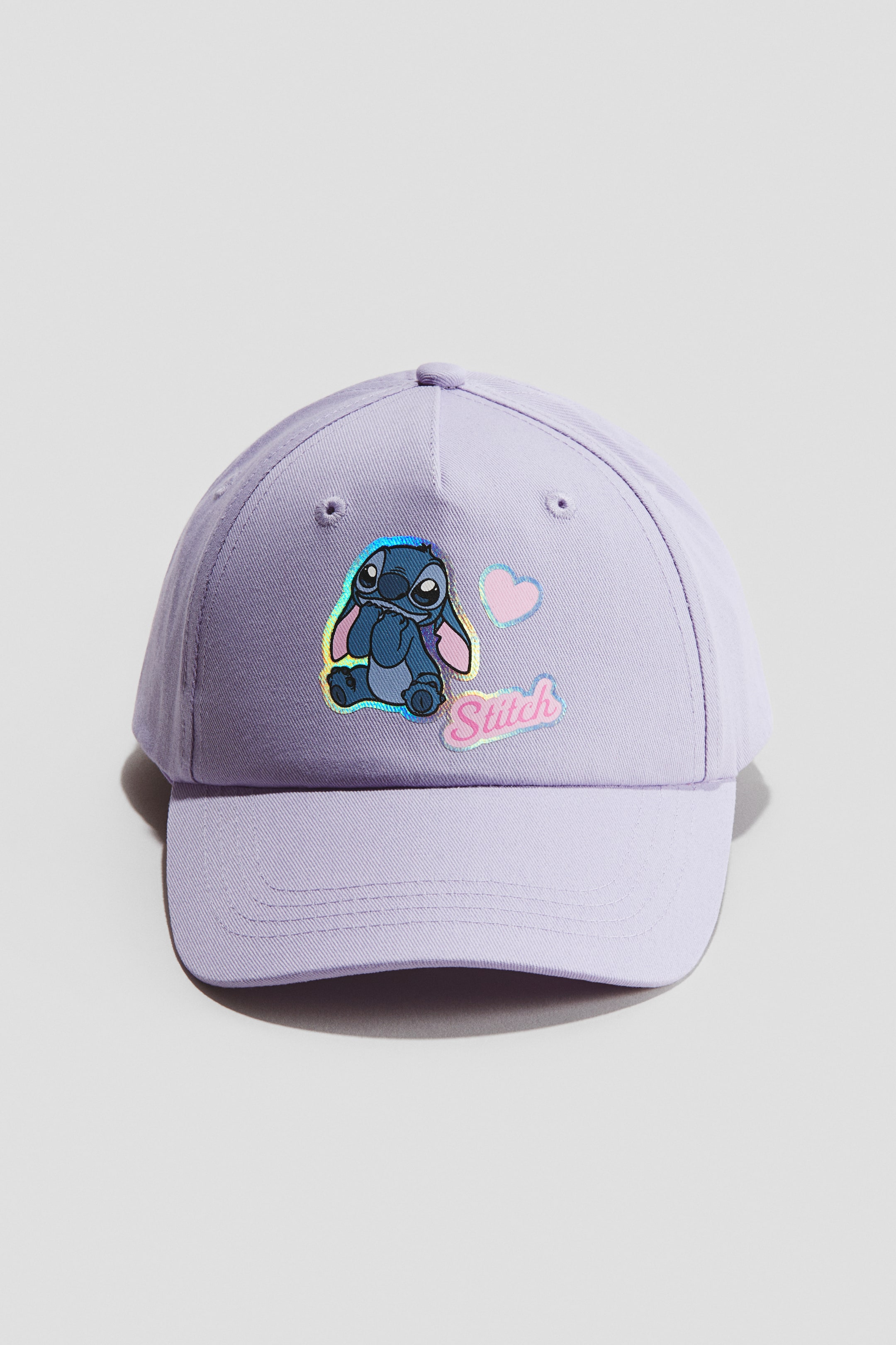 Ver imagen más grande: Una gorra de color púrpura claro en sarga de algodón suave se presenta desde el frente, con una visera curva, una corona de seis paneles con un botón superior y ojales de ventilación bordados. Un gráfico iridiscente del personaje Stitch aparece en el panel frontal, acompañado de un corazón rosa y la palabra 'Stitch' en letras rosas.
