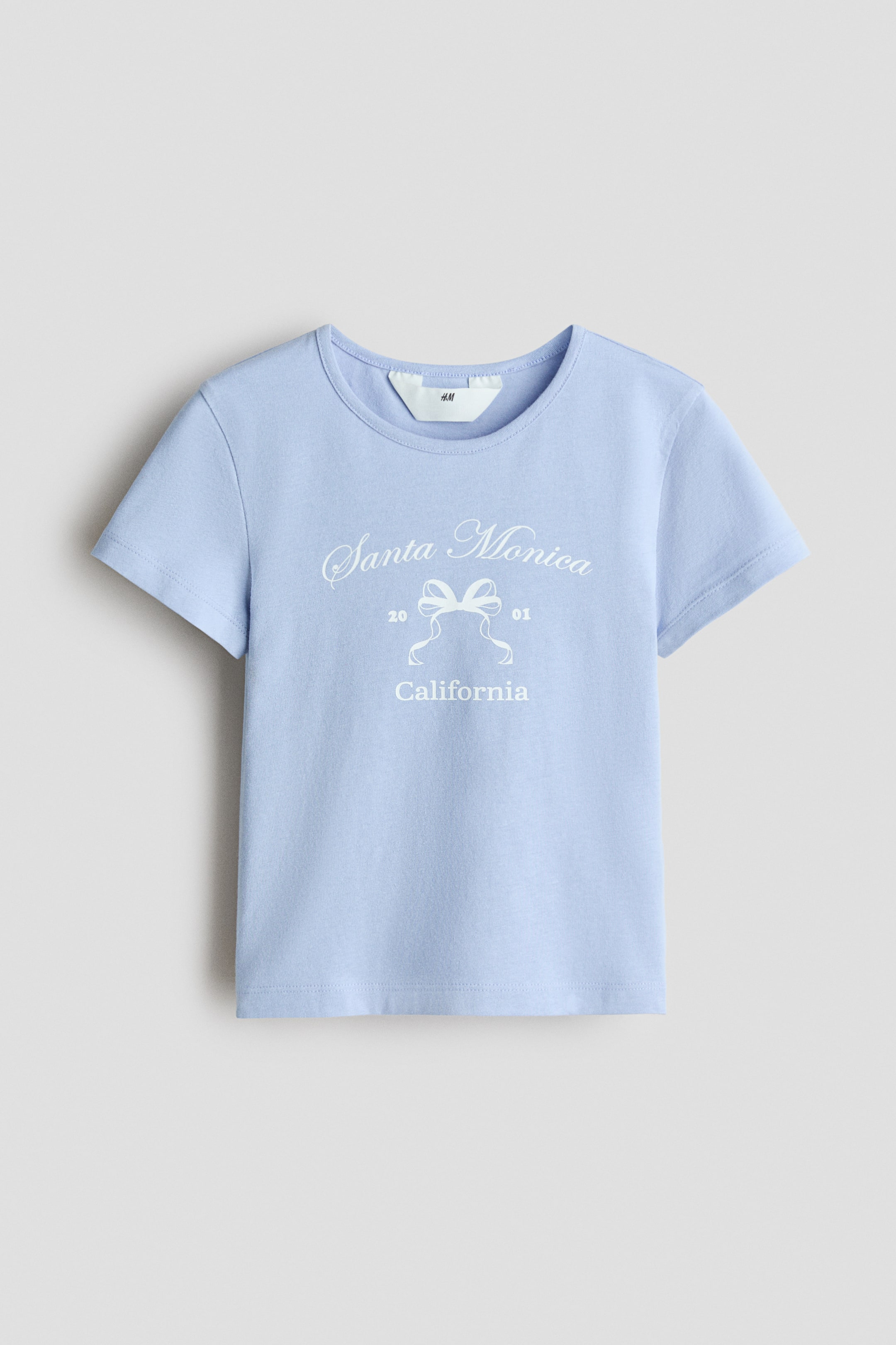 View larger image: A light blue short-sleeved jersey top is presented FRONT, featuring a round neckline. It is displaying a white print motif on the front, which includes "Santa Monica," a bow design, "20 01," and "California."