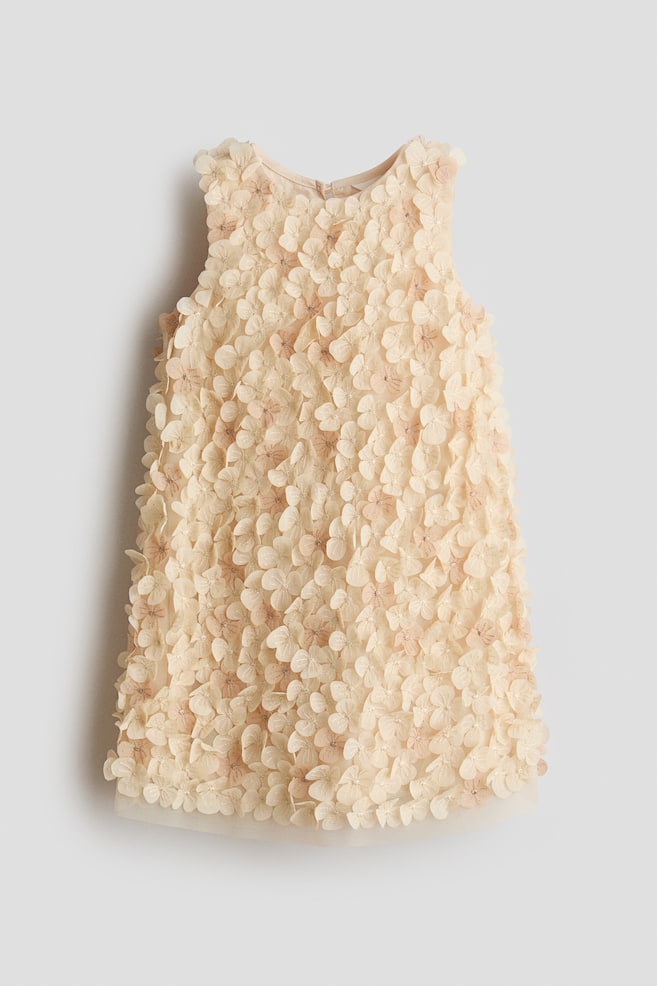 A sleeveless, A-line childrenâs dress is shown from the front, featuring an all-over design of three-dimensional fabric flower appliquÃ©s in cream and light peach shades. The dress has a round neckline and a sheer tulle underlayer peeking out slightly at the hem. - 1