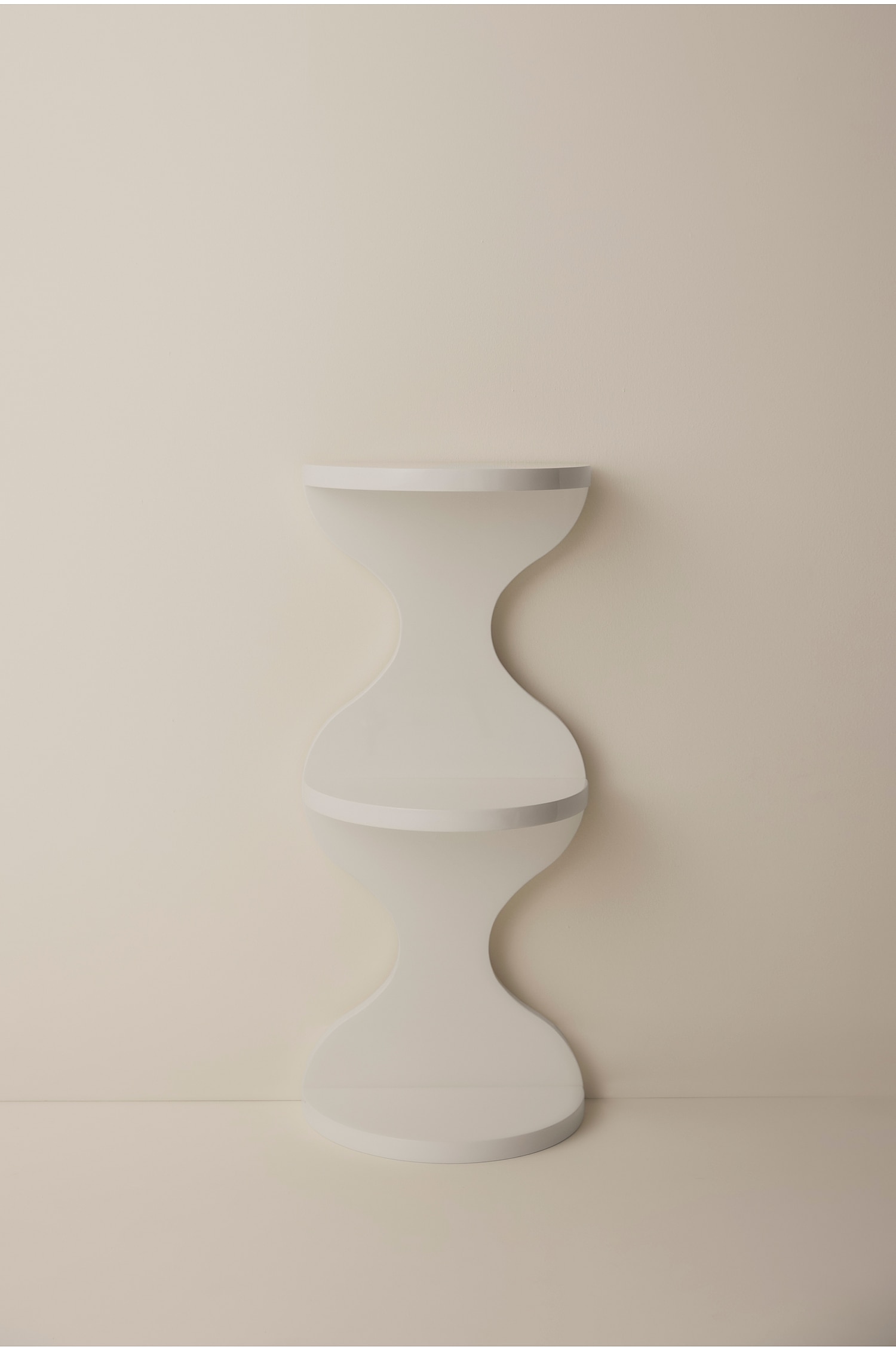 Een lichtbeige sculpturale sokkel staat rechtop en heeft een dubbel zandlopersilhouet met drie ronde niveaus. Het stuk heeft een gladde, matte afwerking met gebogen, symmetrische kolommen die de vlakke basis, de middelste plank en het bovenste oppervlak met elkaar verbinden.