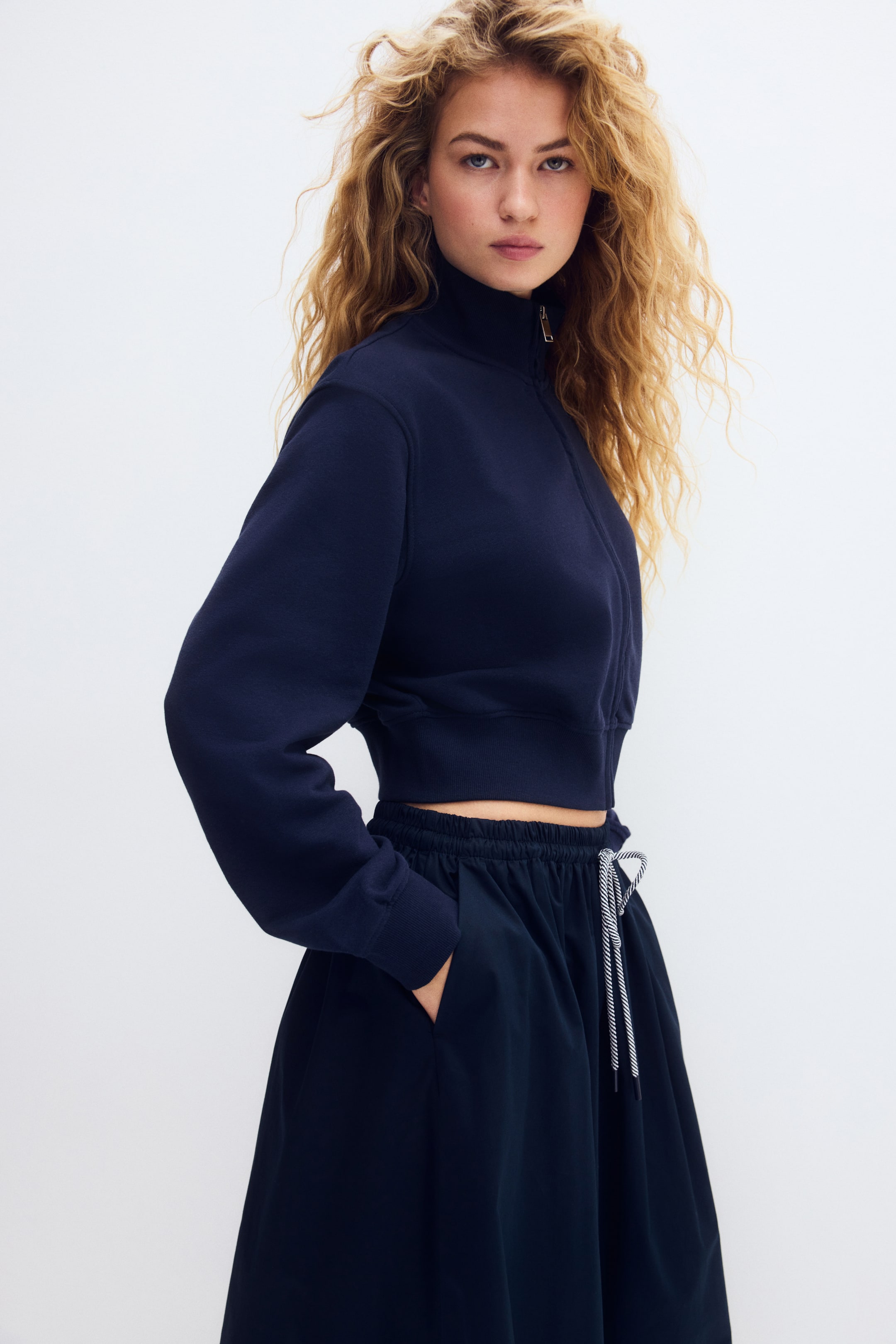 View larger image: A navy blue cropped sweatshirt features a high collar with a silver-toned quarter-zip closure, long sleeves and a ribbed hem. It is styled with a navy blue A-line skirt featuring an elasticated waistband and a white-and-navy striped drawstring.