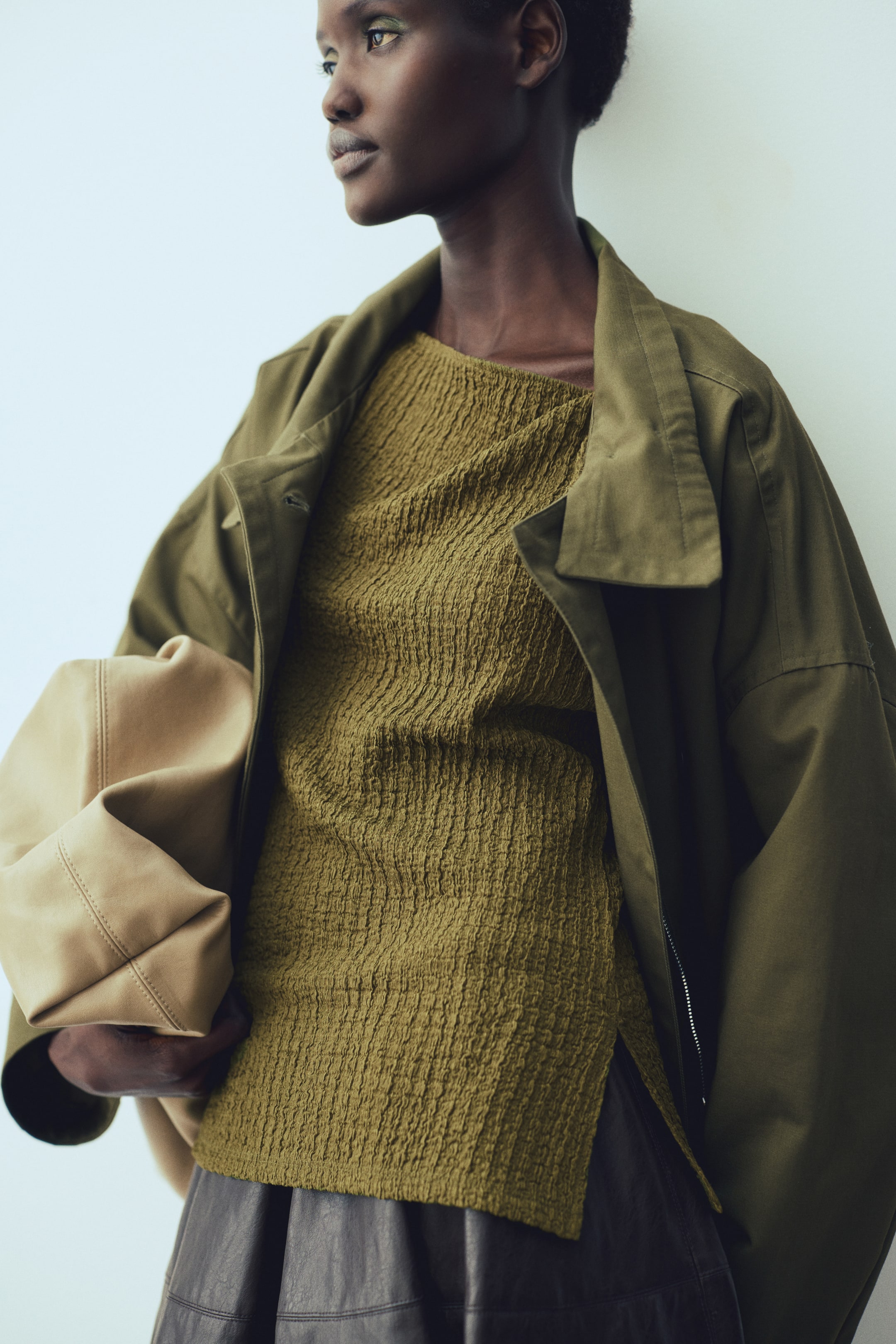 Xem ảnh lớn hơn: Người mẫu đang mặc một áo ôm sát màu xanh ô-liu bằng jersey có kết cấu, với cổ áo bất đối xứng và hiệu ứng drap nhẹ nhàng. Người mẫu khoác thêm một chiếc áo khoác màu xanh bên ngoài, mặc một chiếc chân váy tối màu và cầm một chiếc túi màu be.