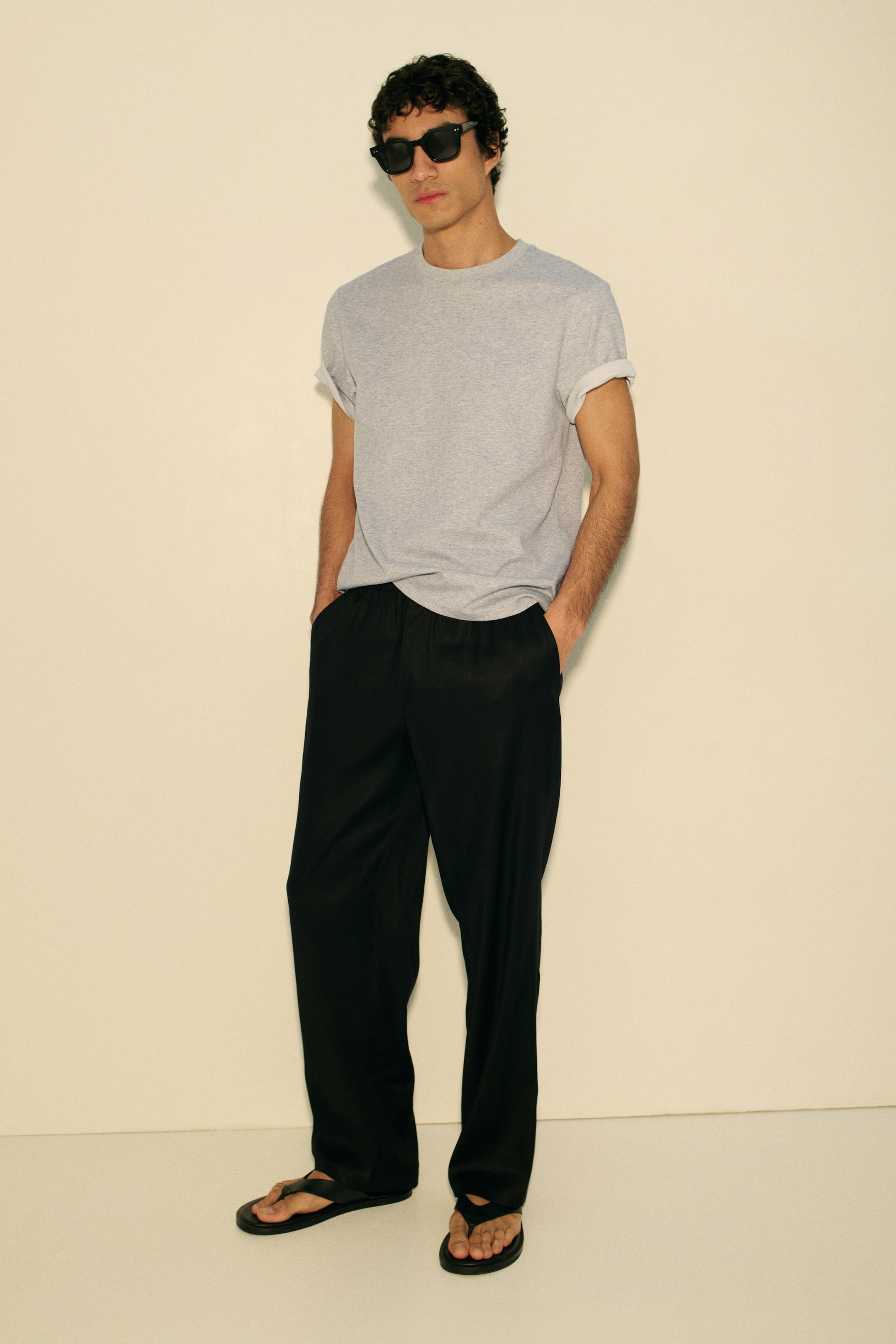 View larger image: A light grey crew neck t-shirt featuring rolled short sleeves and a relaxed fit, teamed with black loose-fit trousers showcasing a wide-leg silhouette and visible side pockets. Black thong-style flip-flops are worn underneath the trousers.