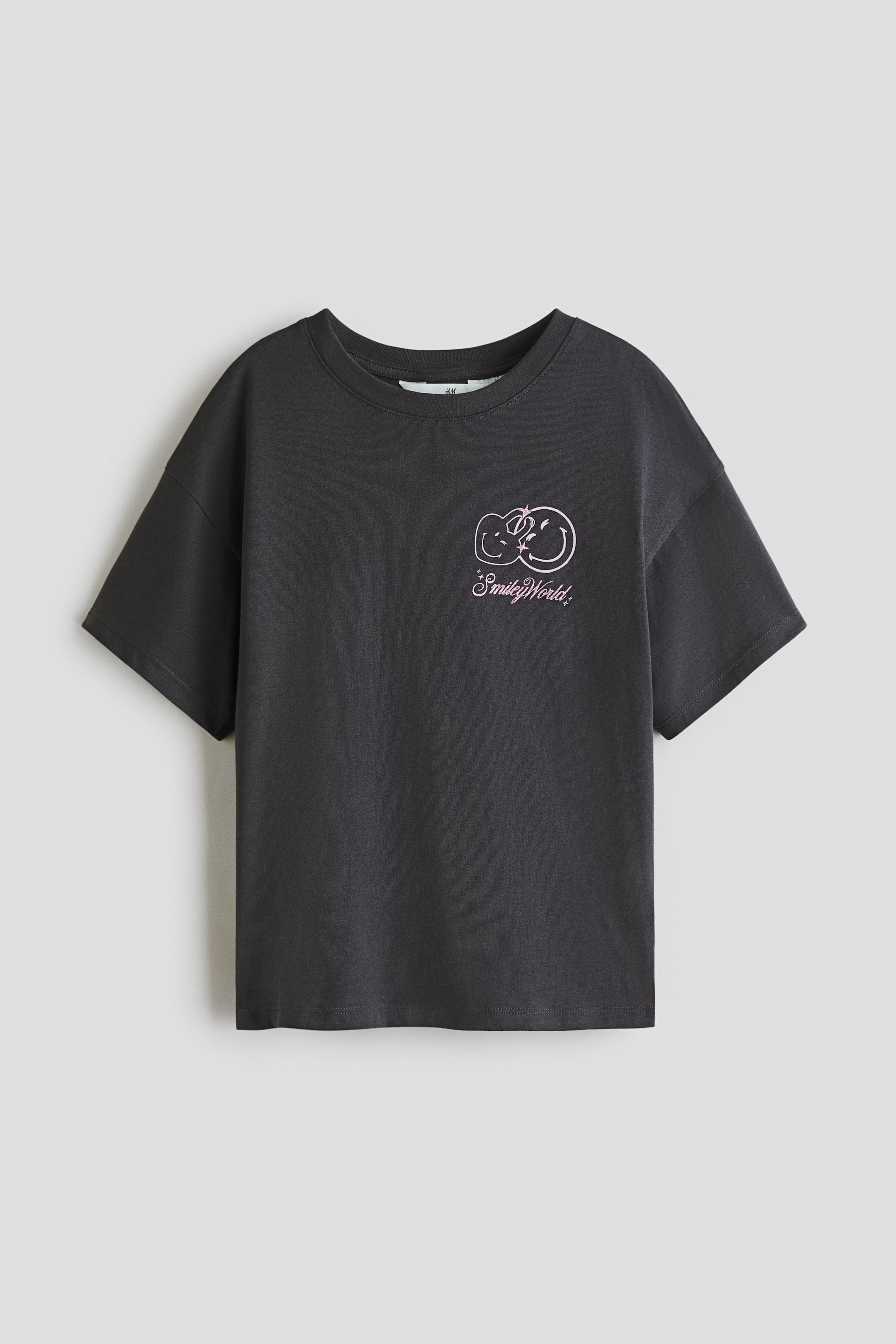 View larger image: A dark grey t-shirt is displayed front-facing, laid flat, showcasing a round neck and short, relaxed-fit sleeves. The front chest features a pink and white graphic with two interlocking smiley faces and the text "SmileyWorld."