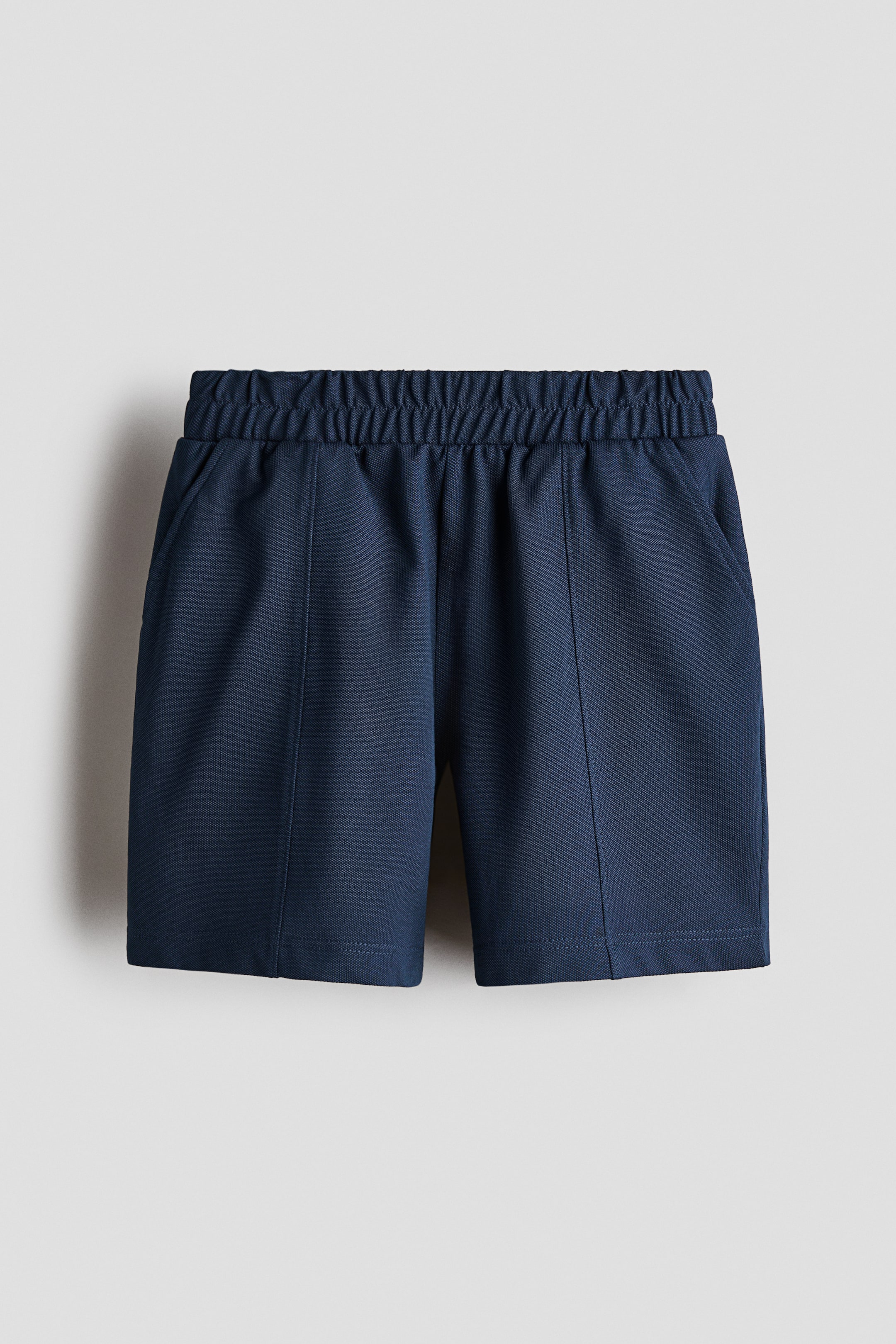 Xem ảnh lớn hơn: Quần short màu xanh navy được chụp ở góc nhìn chính diện, với cạp chun nhún và túi hai bên. Các đường may dọc chạy xuống giữa mỗi ống quần, kết thúc bằng lai gấp đôi được may kép.