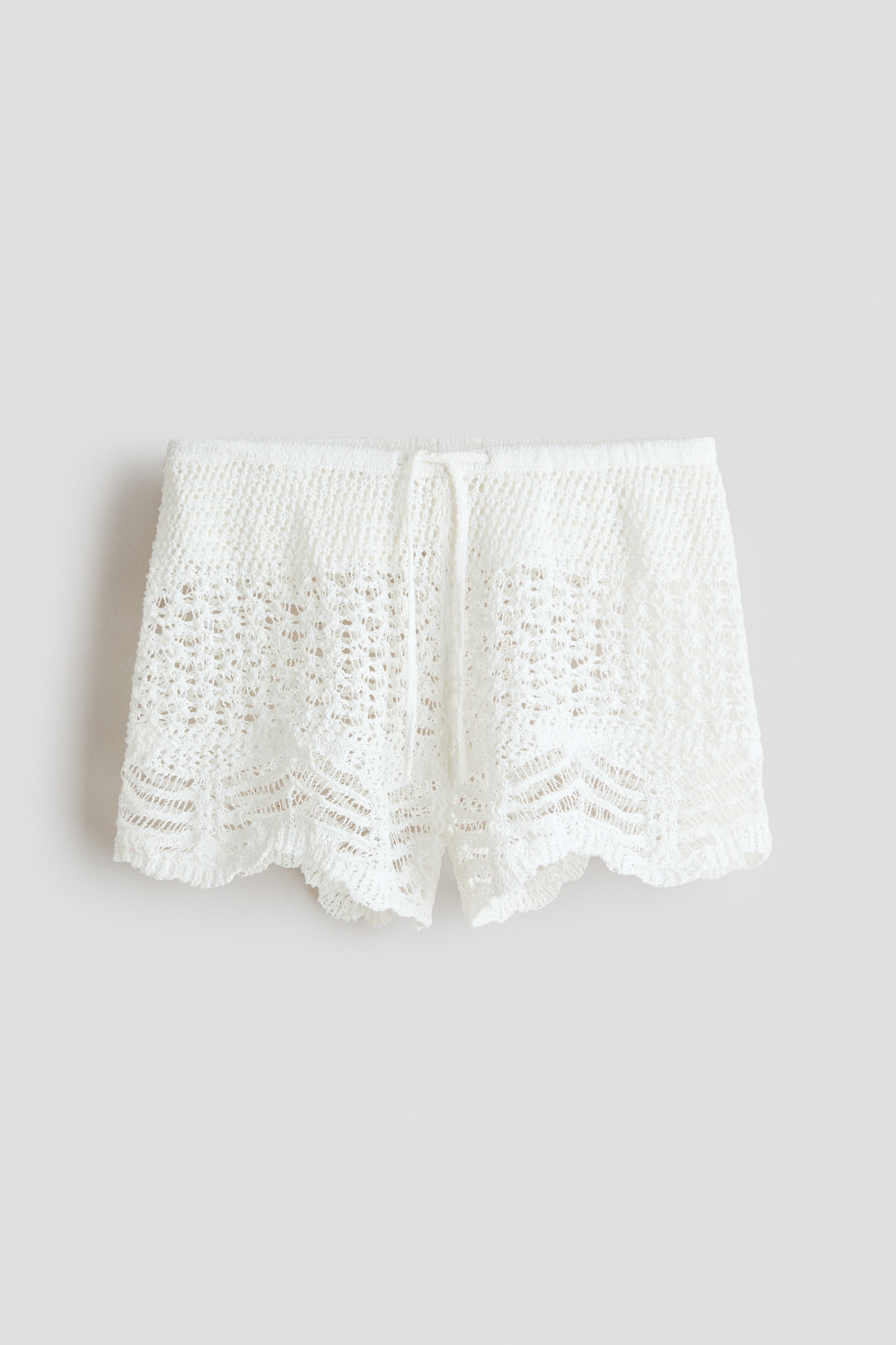 Agrandir l'image: Les shorts en crochet blanc présentent un motif délicat en maille ajourée. Ils sont dotés d'une taille élastiquée avec un cordon de serrage central et des ouvertures de jambes festonnées.