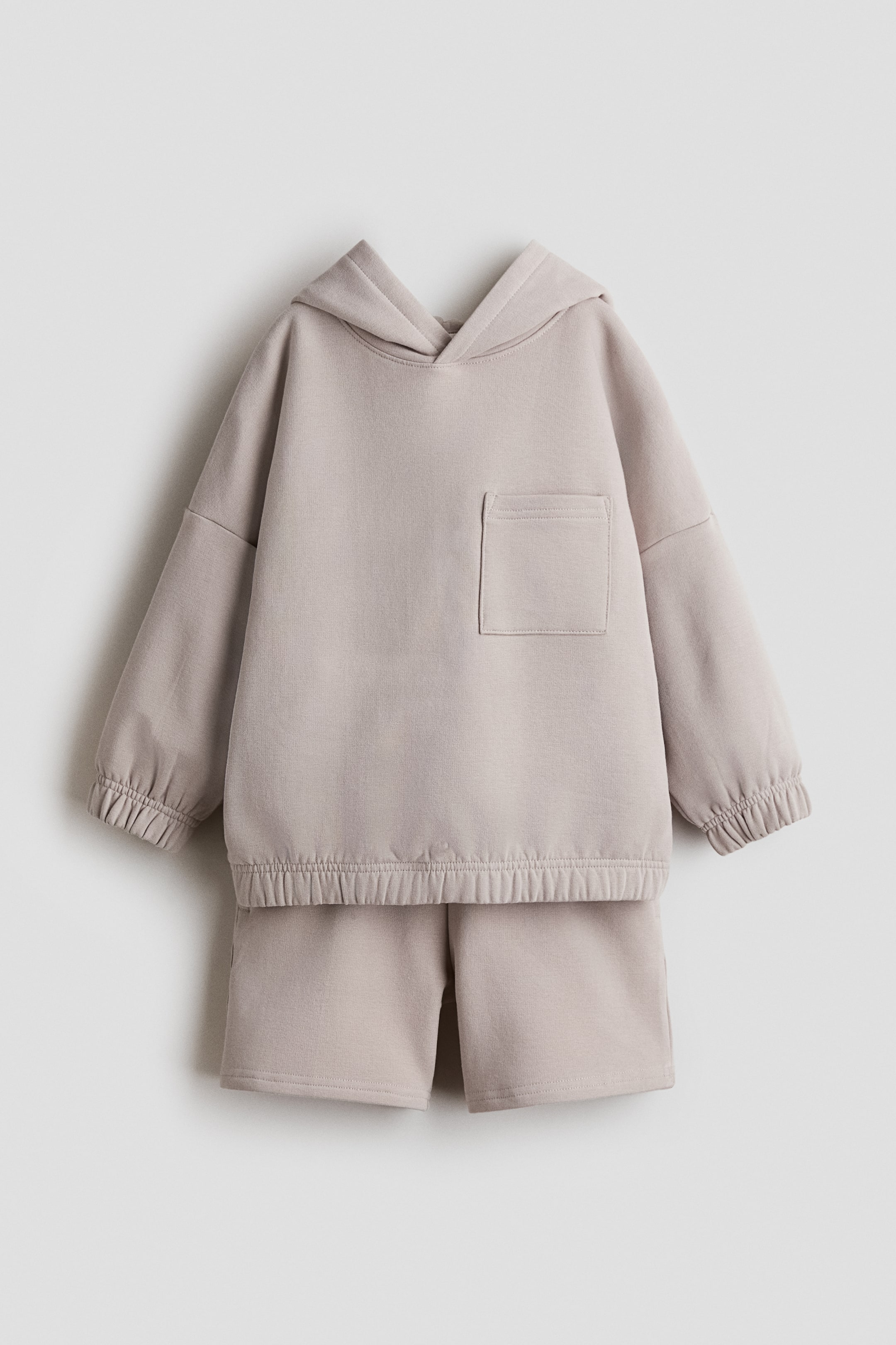 Se større billede: Et lysebeige todelt loungewear-sæt præsenteres forfra. Sættet inkluderer en hættetrøje med hætte, en brystlomme på venstre side og elastiske manchetter og kant, samt ensfarvede matchende shorts.